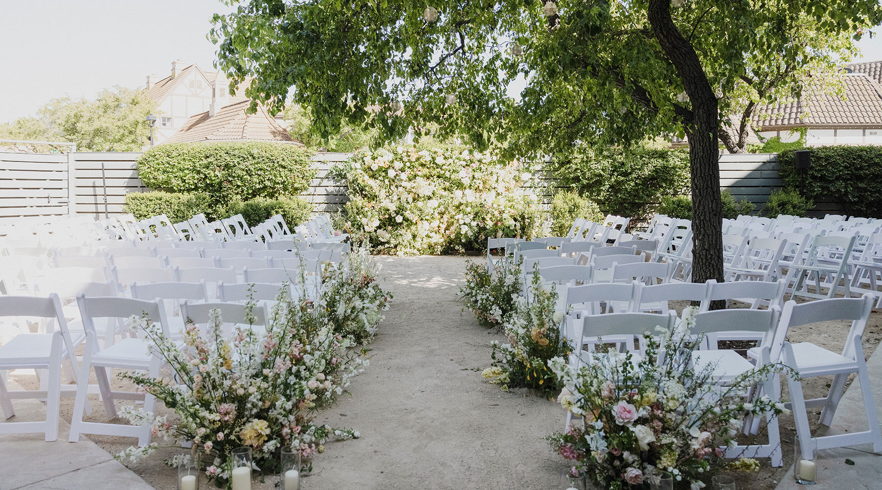 Outdoor wedding ceremony setup with rows of white chairs facing a floral arch, greenery and blooming flowers lining the aisle, and a tree providing shade in a sunny garden setting.