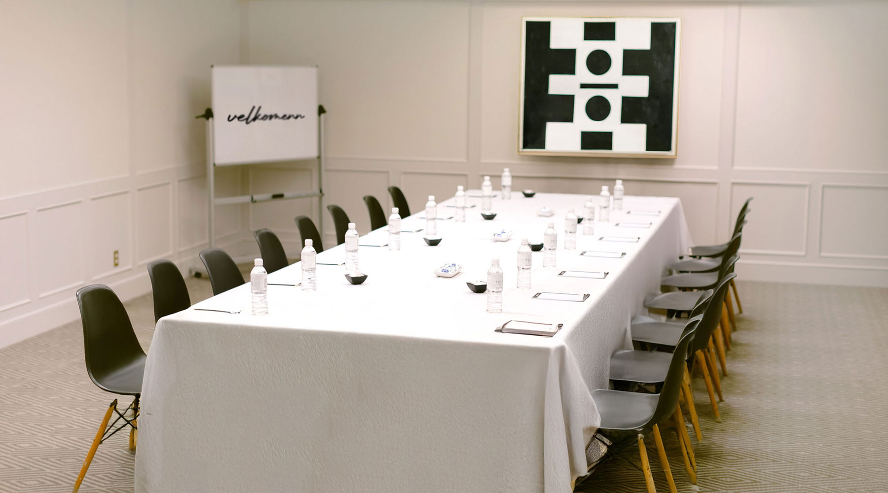 A conference room with a long, white-covered table set for a meeting. Black chairs surround the table, which has bottled water, notepads, and bowls. A whiteboard with welcome and abstract art are in the background.