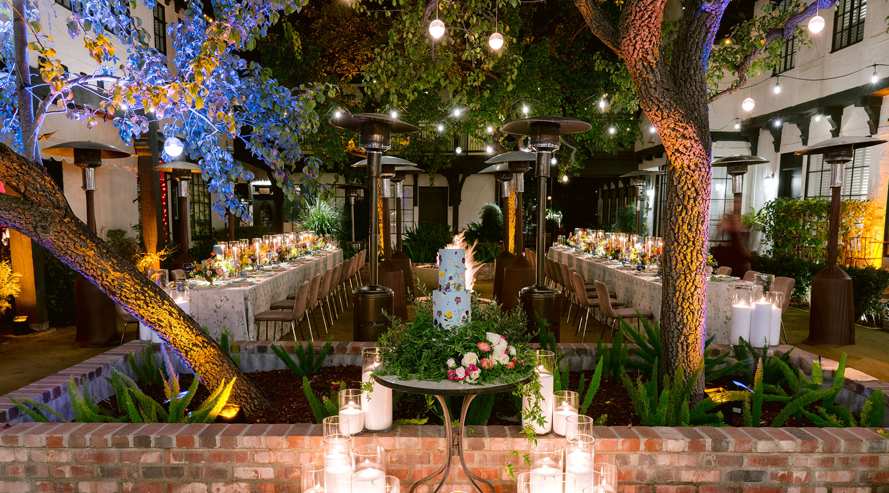 An outdoor garden wedding reception at night, featuring long tables with floral centerpieces, string lights overhead, patio heaters, and a decorated cake on a central table surrounded by candles and greenery.