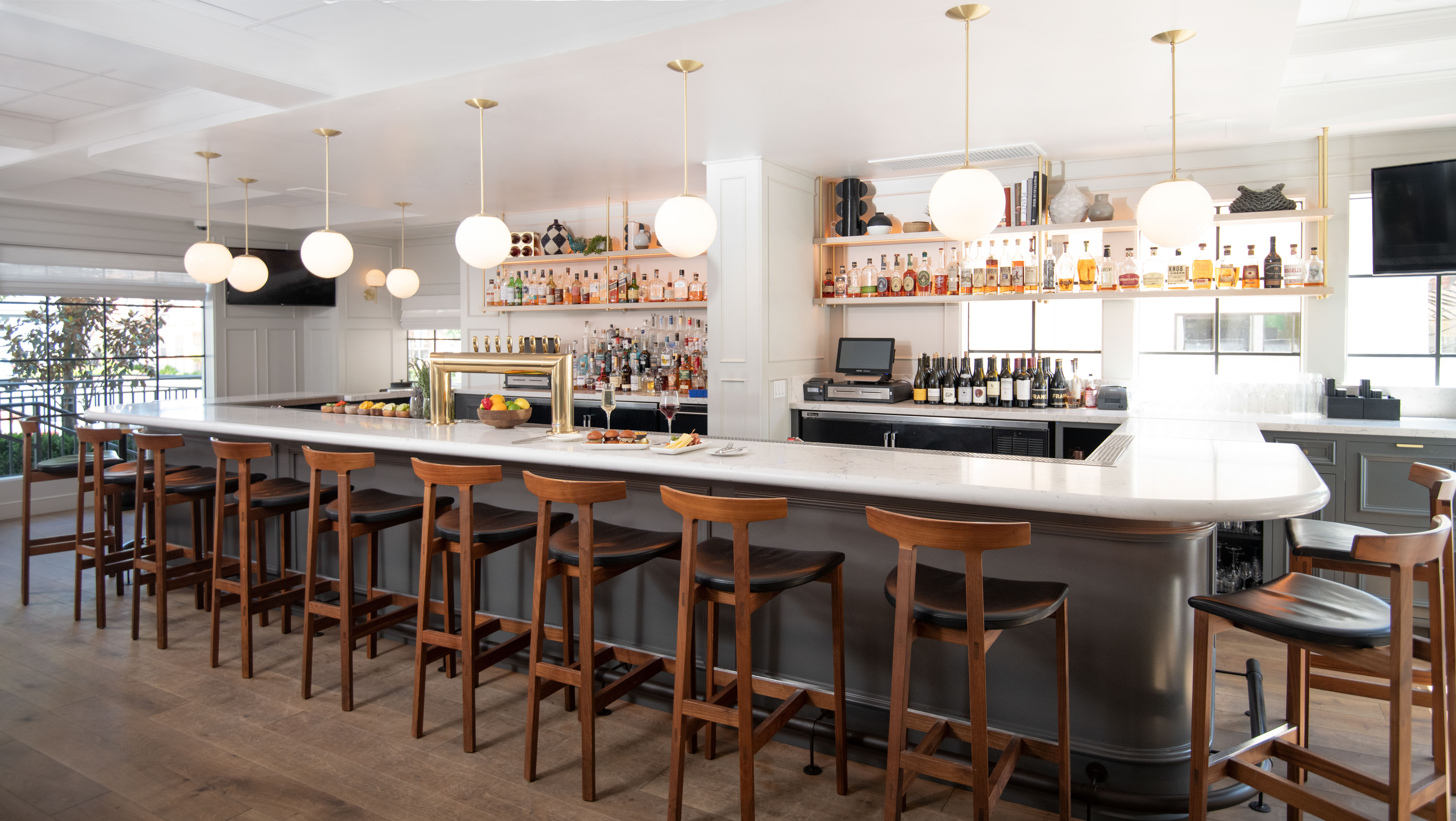 Photo of The Landsby. A modern bar with a long white countertop, wooden bar stools, hanging globe lights, and shelves stocked with bottles of liquor and wine. The bar is empty, with natural light coming in through large windows.