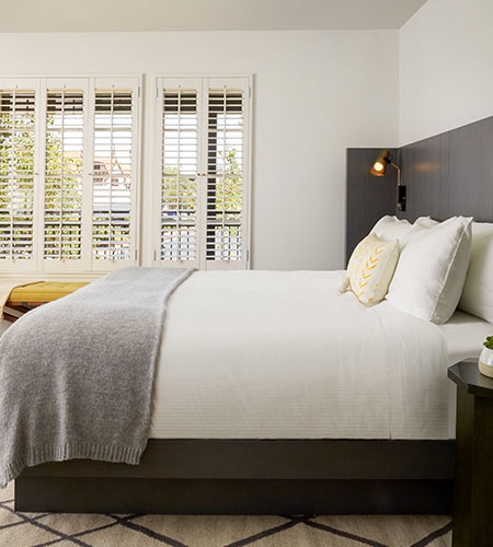 A modern bedroom with a neatly made bed featuring white linens, gray blanket, and decorative pillows. Large windows with white shutters let in natural light. A small bedside table and lamp are visible.