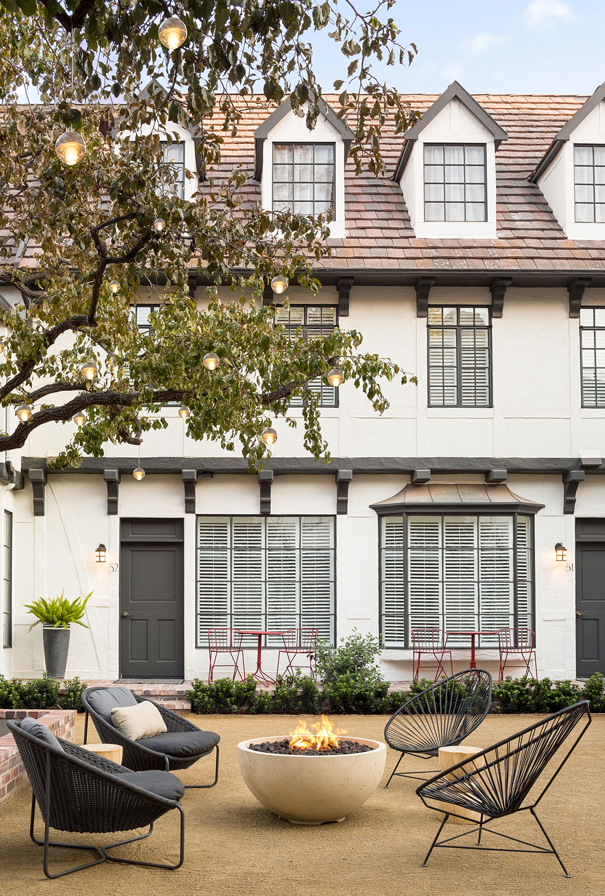 Photo of The Landsby. A cozy courtyard with modern black chairs around a round fire pit, string lights overhead, and a white townhouse with gray trim and red bistro tables in the background.