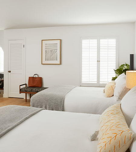 A bright, modern bedroom with two neatly made beds, white walls, a window with shutters, a bench with a brown bag, framed art, and a small plant in the corner.