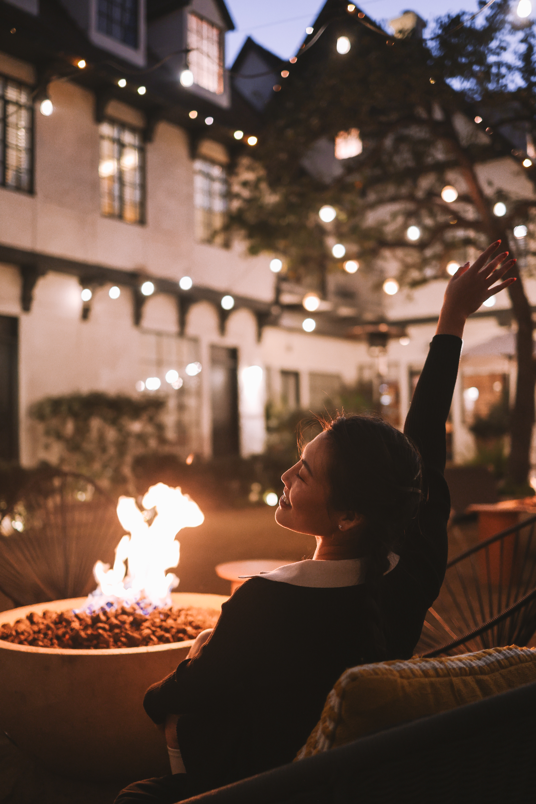 Photo of The Landsby. A woman smiles and raises her arm while sitting by a fire pit outdoors in the evening, surrounded by string lights and a building in the background.