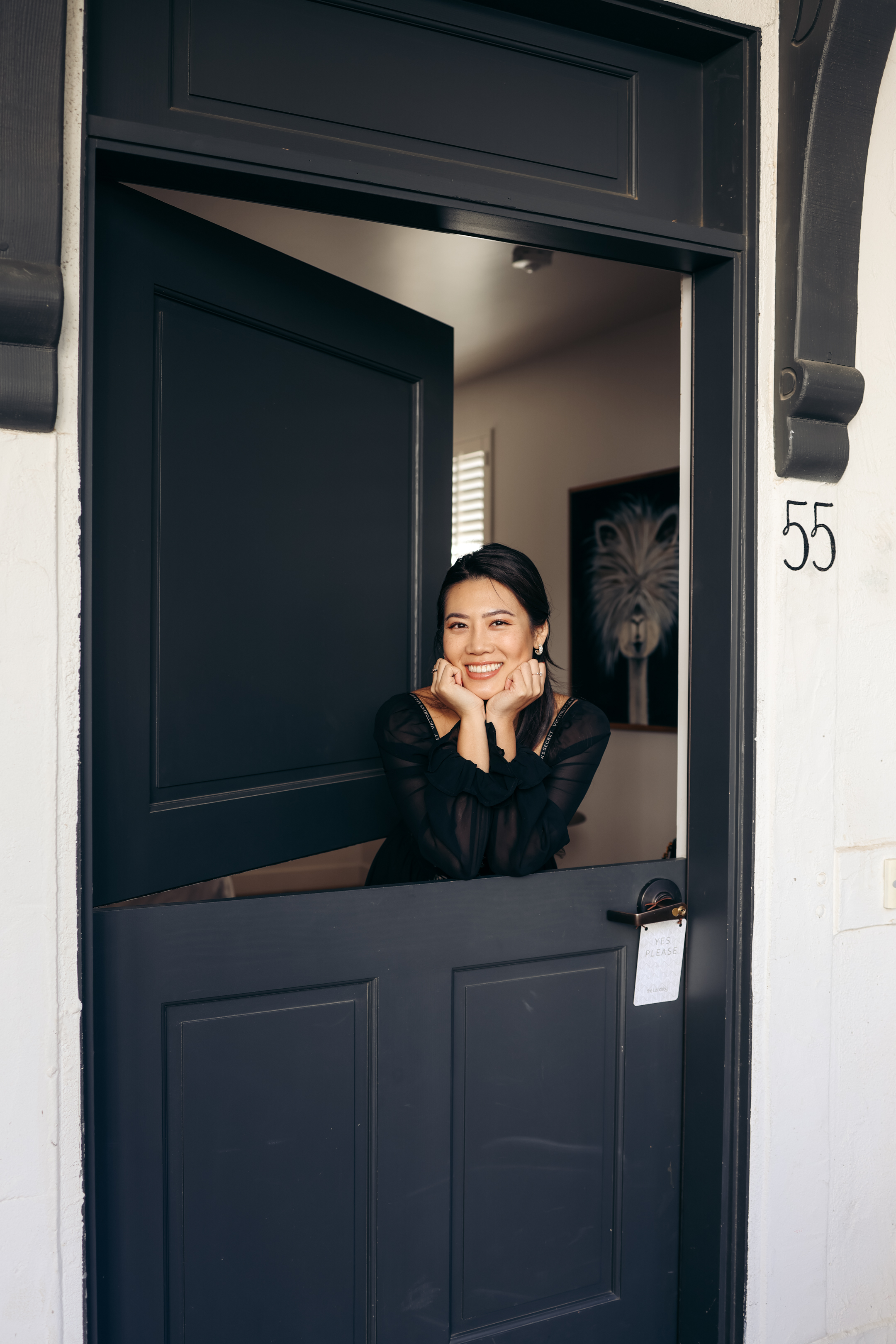 Photo of The Landsby. A woman in a black top smiles and leans on the lower half of an open black Dutch door, with her elbows resting on the door. The door has the number 55, and a room is visible in the background.