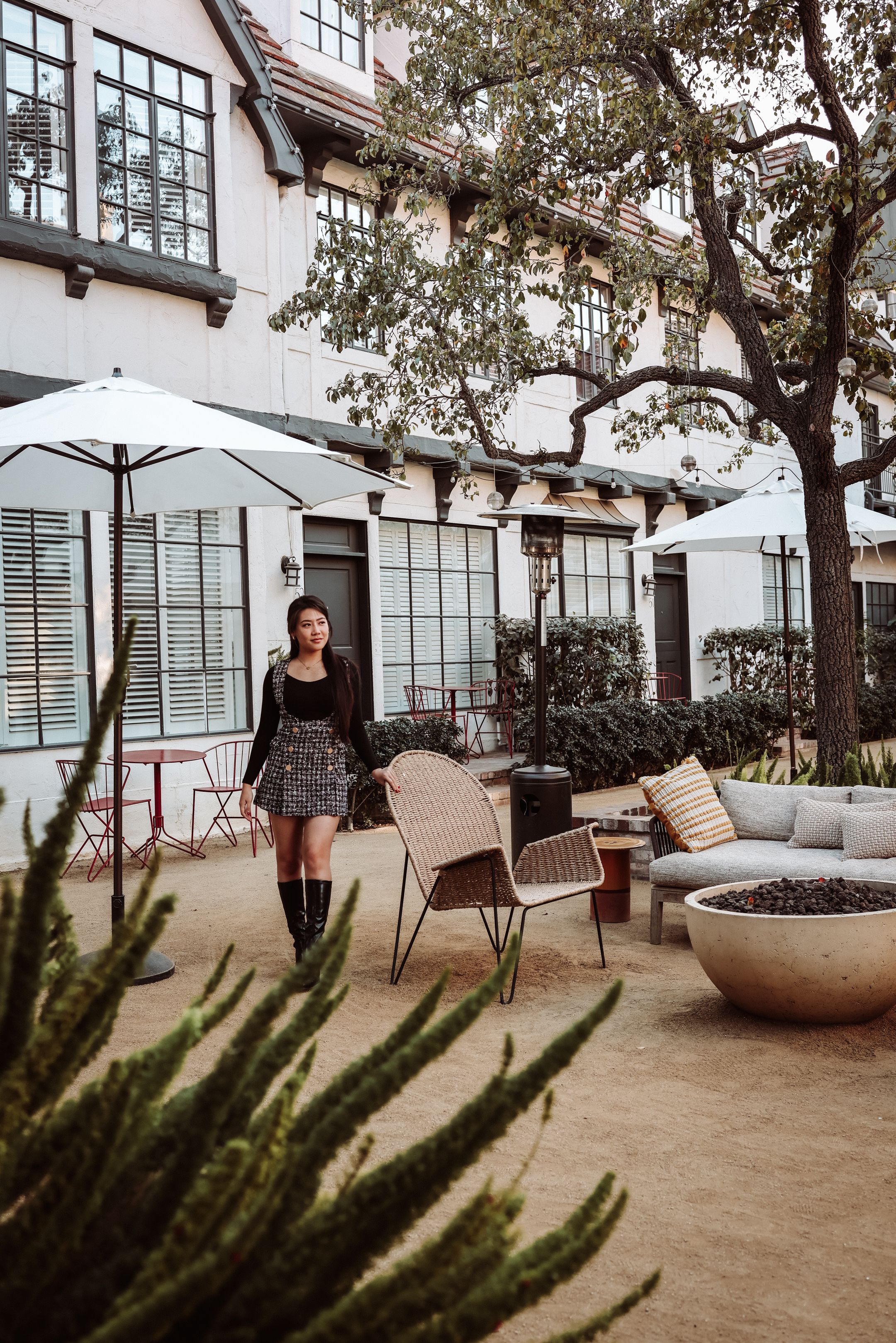 Photo of The Landsby. A woman in a black top, plaid skirt, and knee-high boots stands in a stylish outdoor patio area with wicker chairs, a fire pit, and large white umbrellas, in front of a building with many windows.