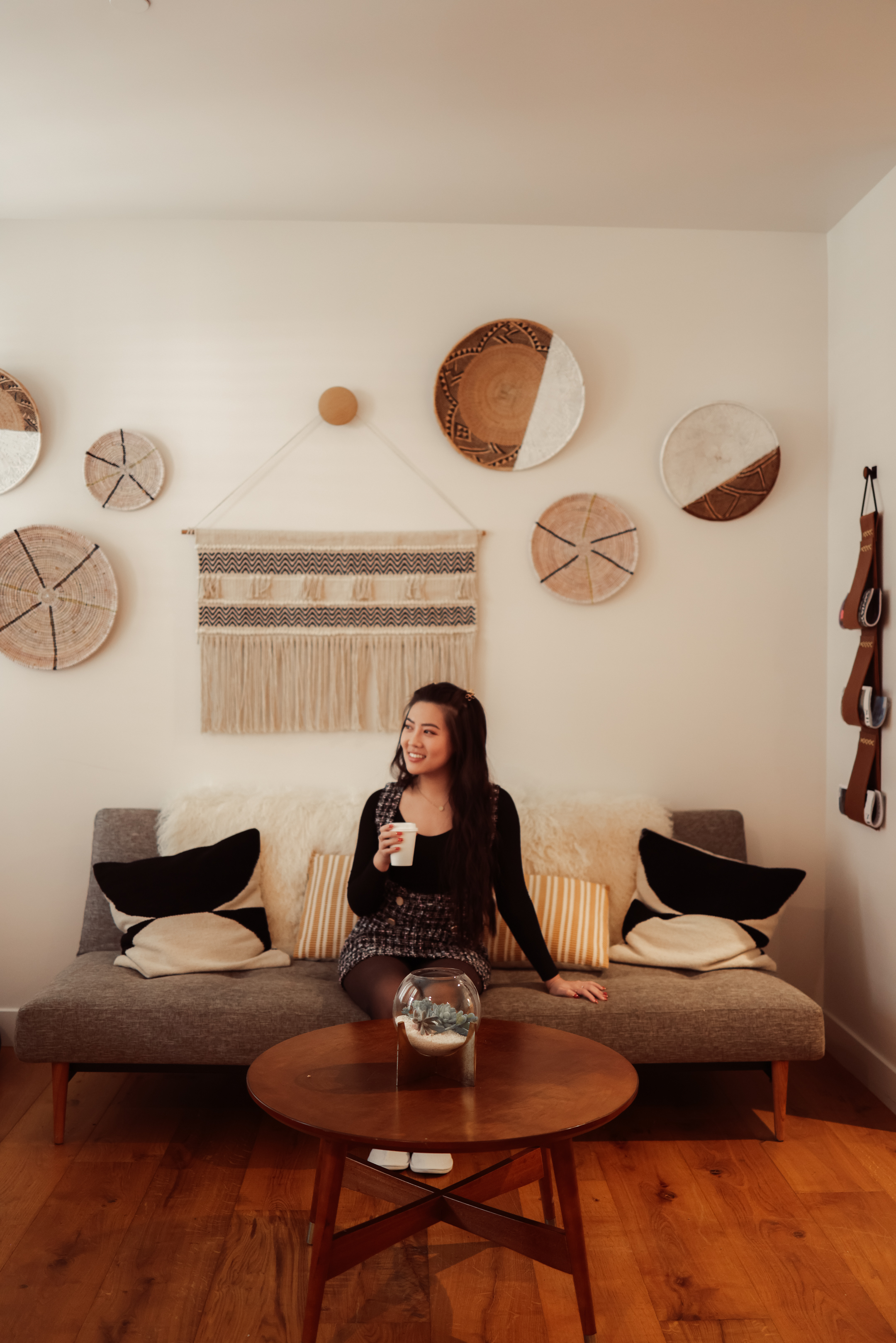 Photo of The Landsby. A woman sits on a gray sofa holding a cup, surrounded by pillows. Behind her, woven wall decor and circular art hang on a white wall. A round wooden table with a glass bowl is in front of the sofa.
