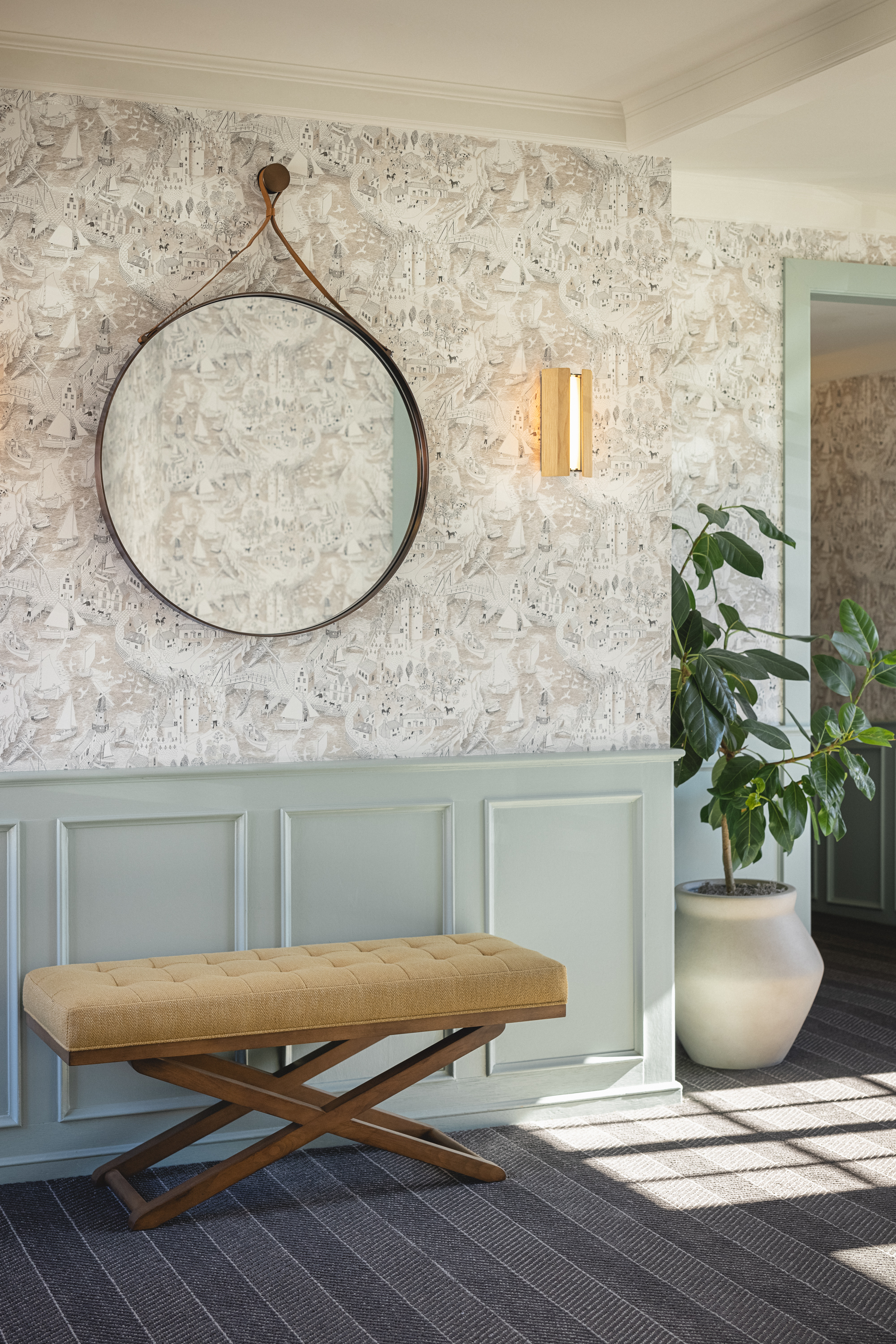 Photo of The Landsby. A round mirror with a leather strap hangs above a yellow cushioned bench with wooden legs. The wall has patterned wallpaper and green wainscoting. A potted plant stands near an open doorway.