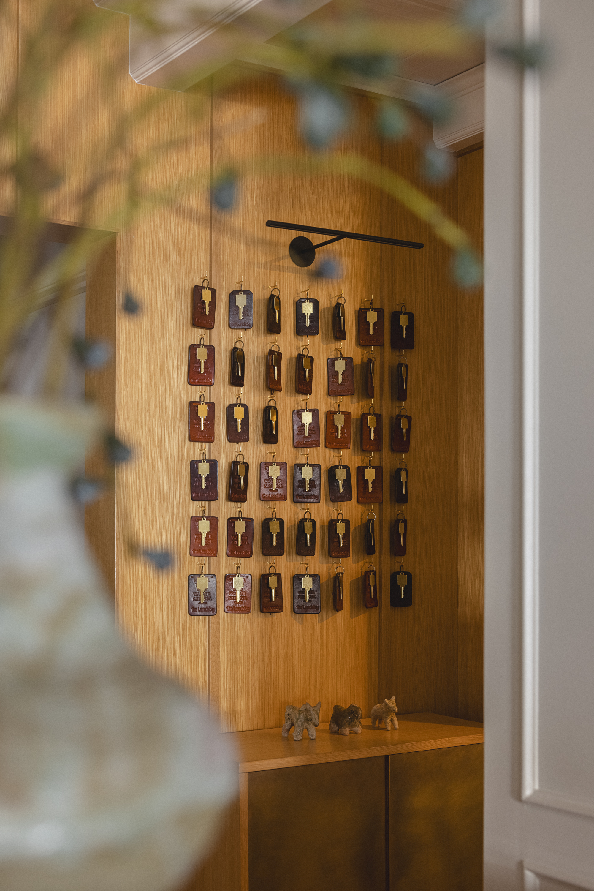 Photo of The Landsby. Wall display of vintage-style leather hotel keychains with brass tags hanging in neat rows on a wooden backdrop. A black modern wall sconce is above, with small decorative animal figurines on a shelf below.