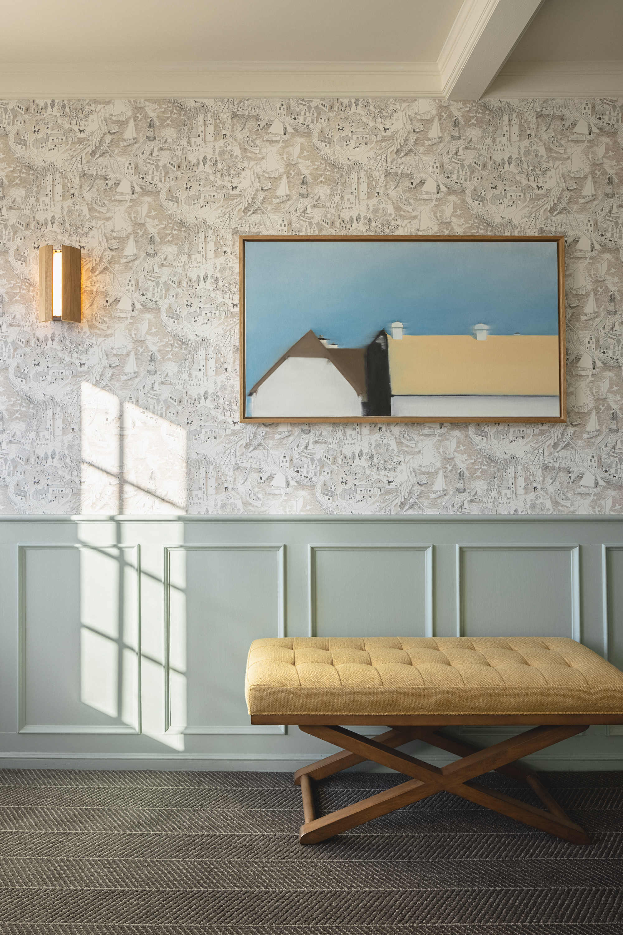 Photo of The Landsby. Interior wall with patterned wallpaper and sage green wainscoting, featuring a framed minimalist painting of rooftops against a blue sky. A mustard-yellow tufted bench with wooden cross legs sits below. Sunlight casts windowpane shadows on the wall.