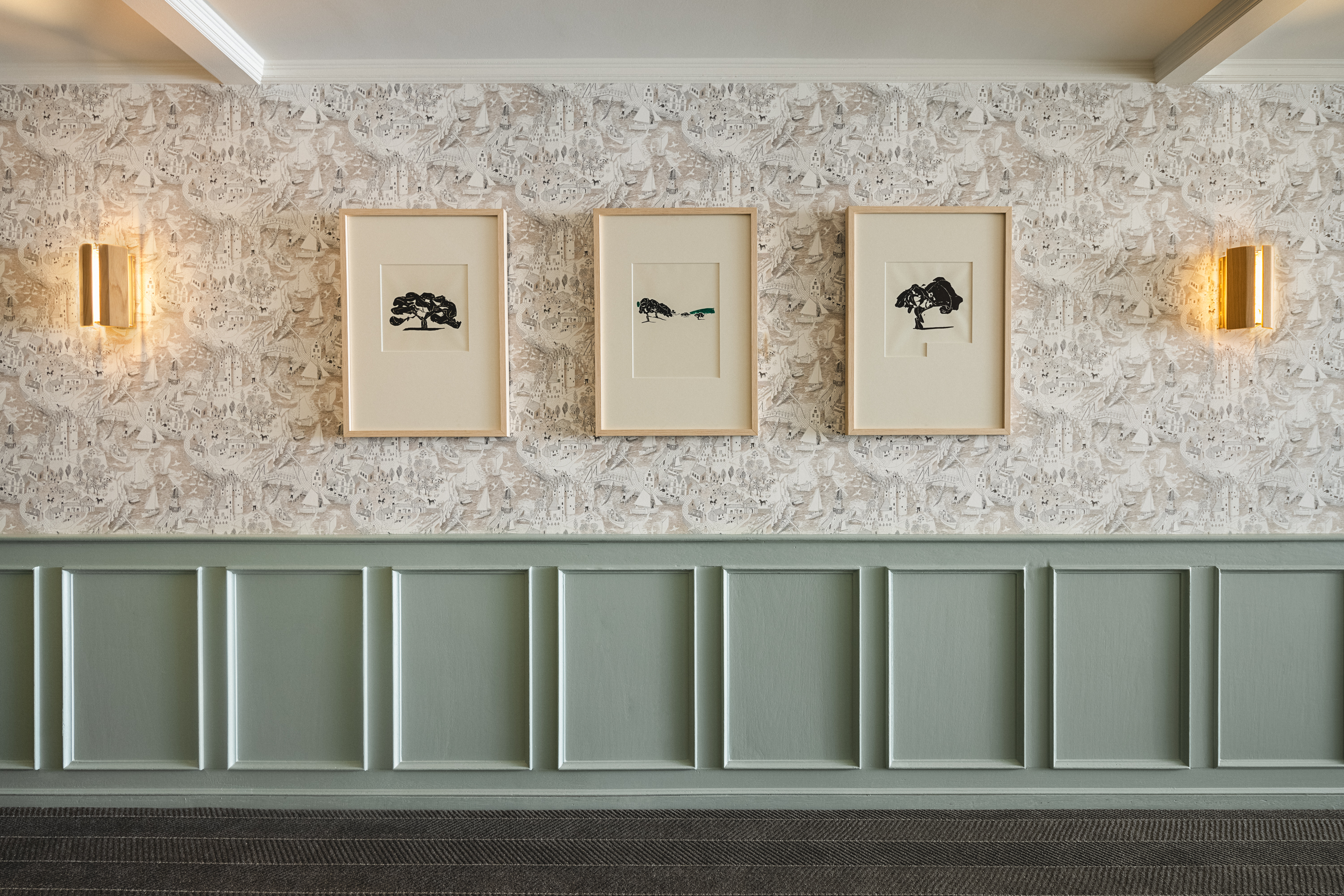 Photo of The Landsby. Interior wall with sage green wainscoting and patterned wallpaper, featuring three framed minimalist tree artworks in a row. Two modern wall sconces provide warm lighting on either side.