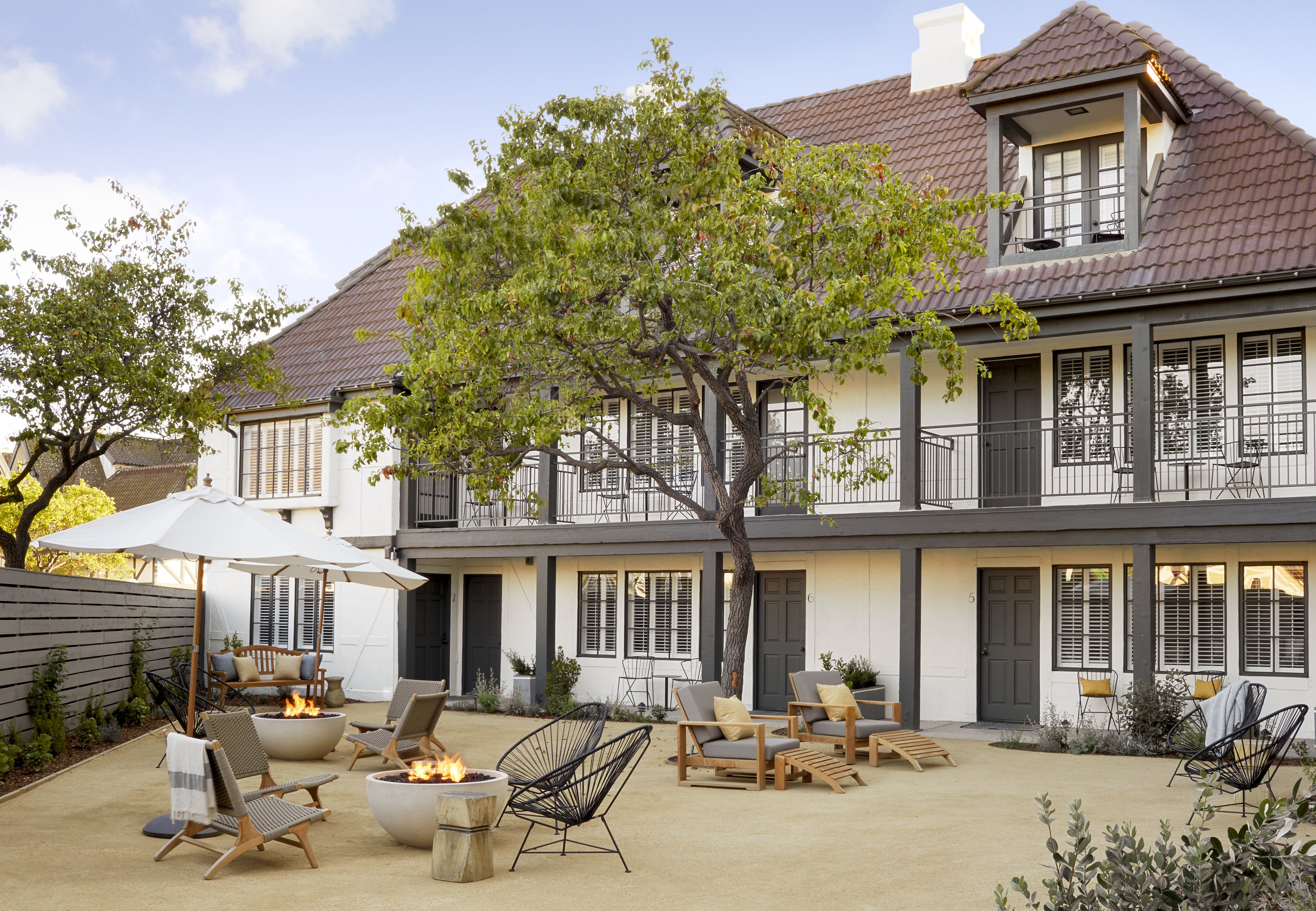 Photo of The Landsby. A courtyard with modern fire pits, lounge chairs, and umbrellas is set in front of a two-story building with a red-tiled roof, grey doors, balconies, and large windows. A leafy tree stands at the center.