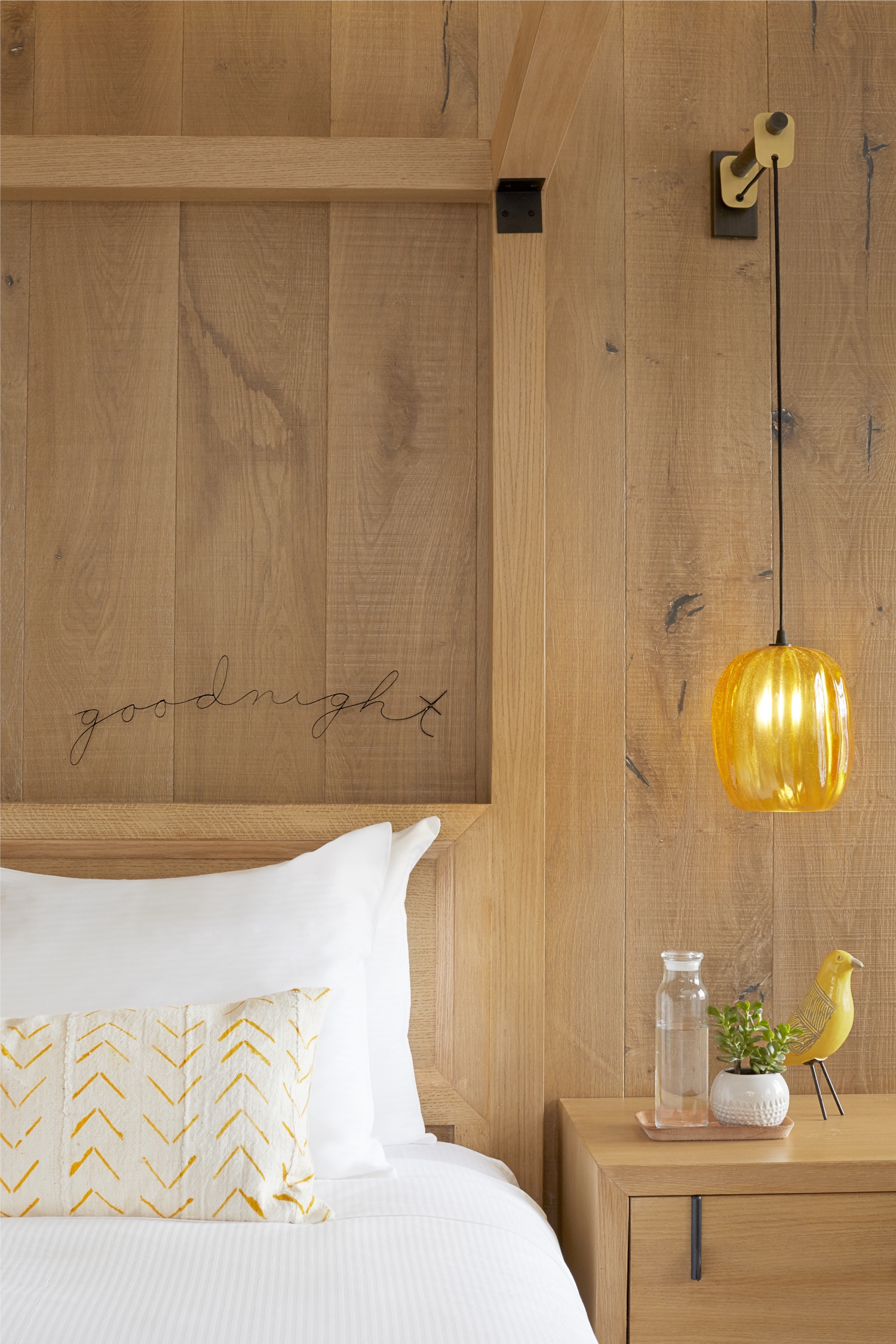 Photo of The Landsby. A cozy bedroom with a wooden headboard reading goodnight, a white bed with a patterned pillow, a wooden nightstand holding a glass of water, small plant, yellow bird figurine, and a hanging amber glass pendant lamp.