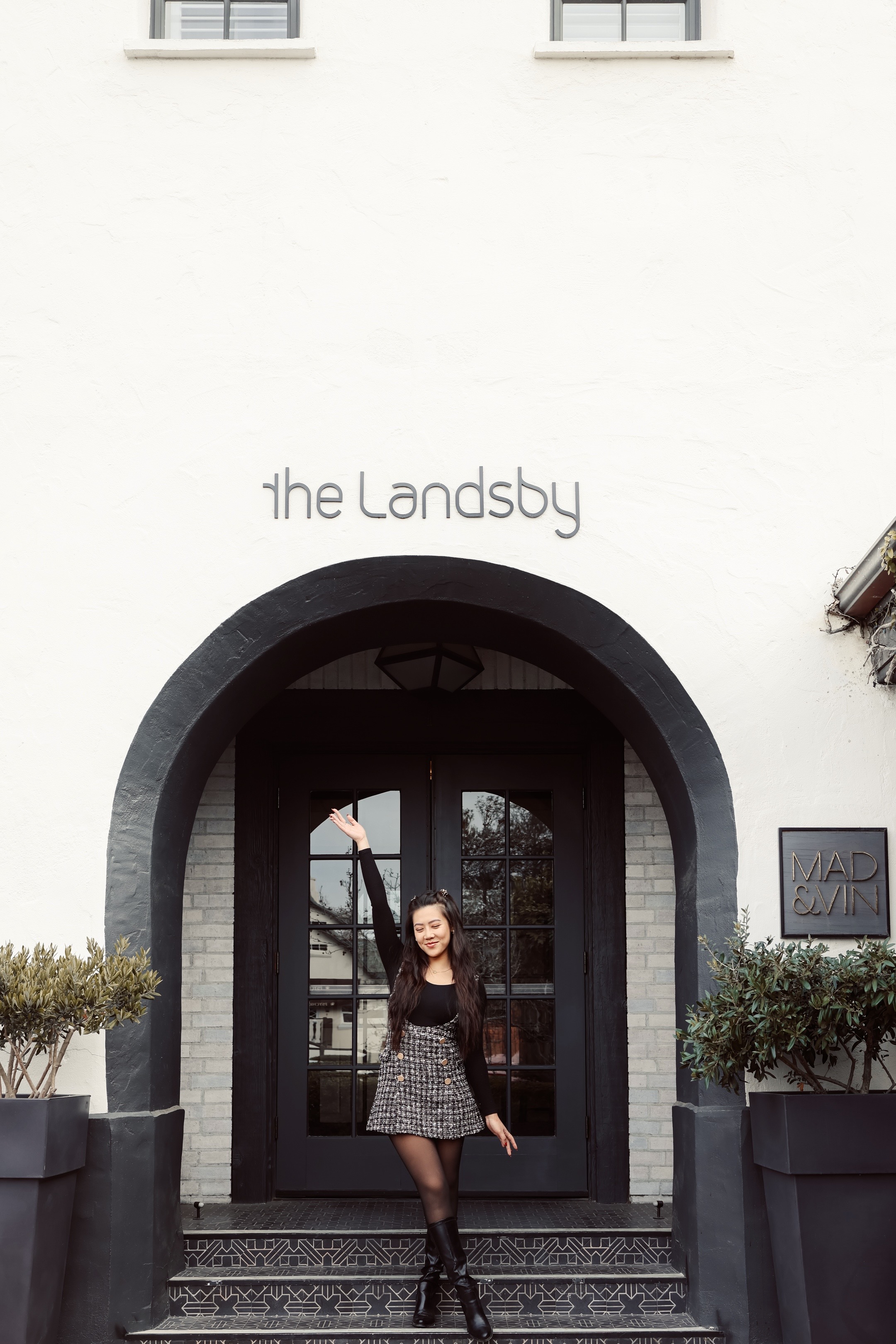 Photo of The Landsby. A woman stands smiling with one arm raised in front of the entrance to The Landsby, a white building with black double doors, potted plants, and a sign reading Mad & Vin to the right.