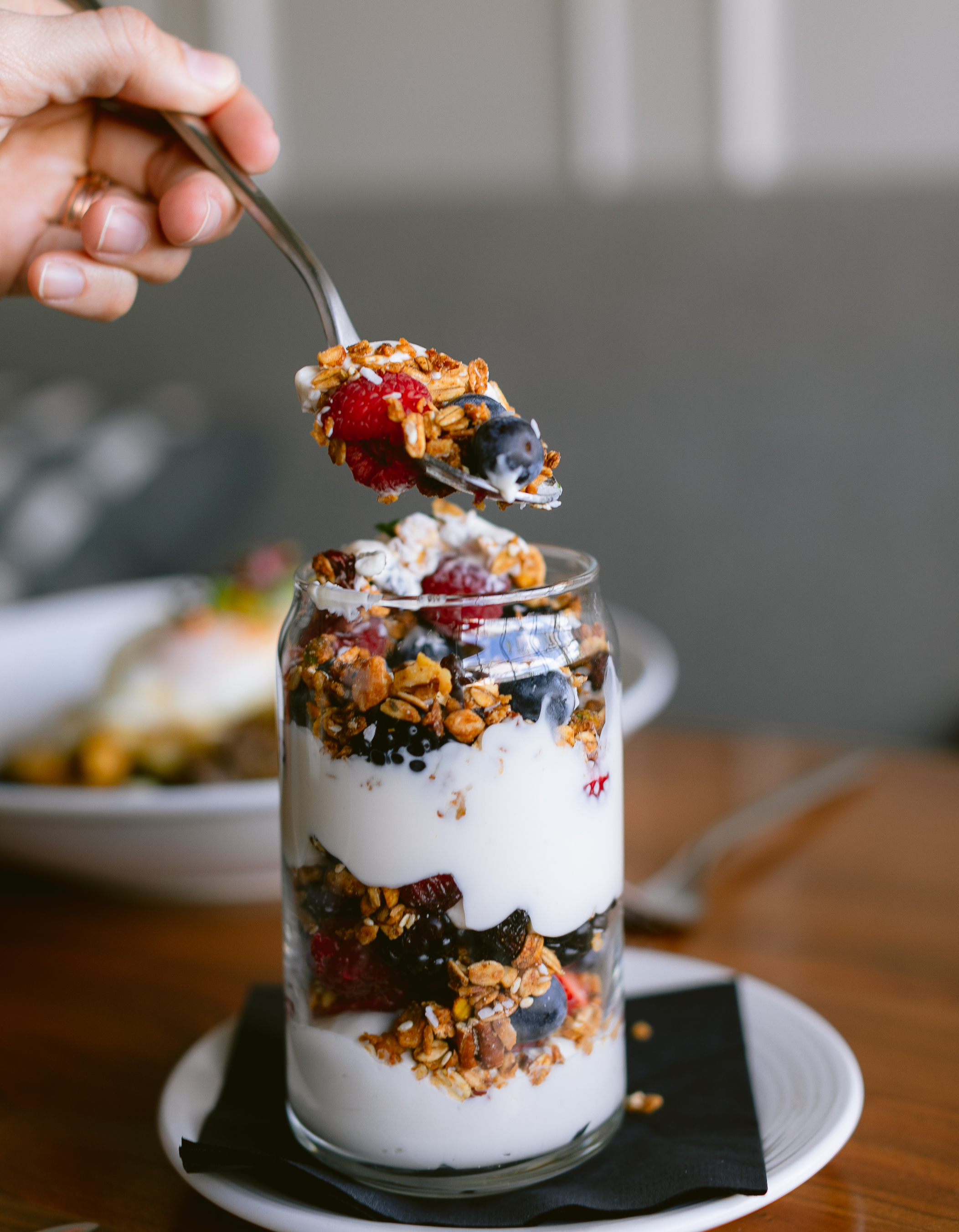 Photo of The Landsby. A hand holding a spoon above a glass filled with layers of yogurt, granola, and mixed berries, with another plate blurred in the background on a wooden table.