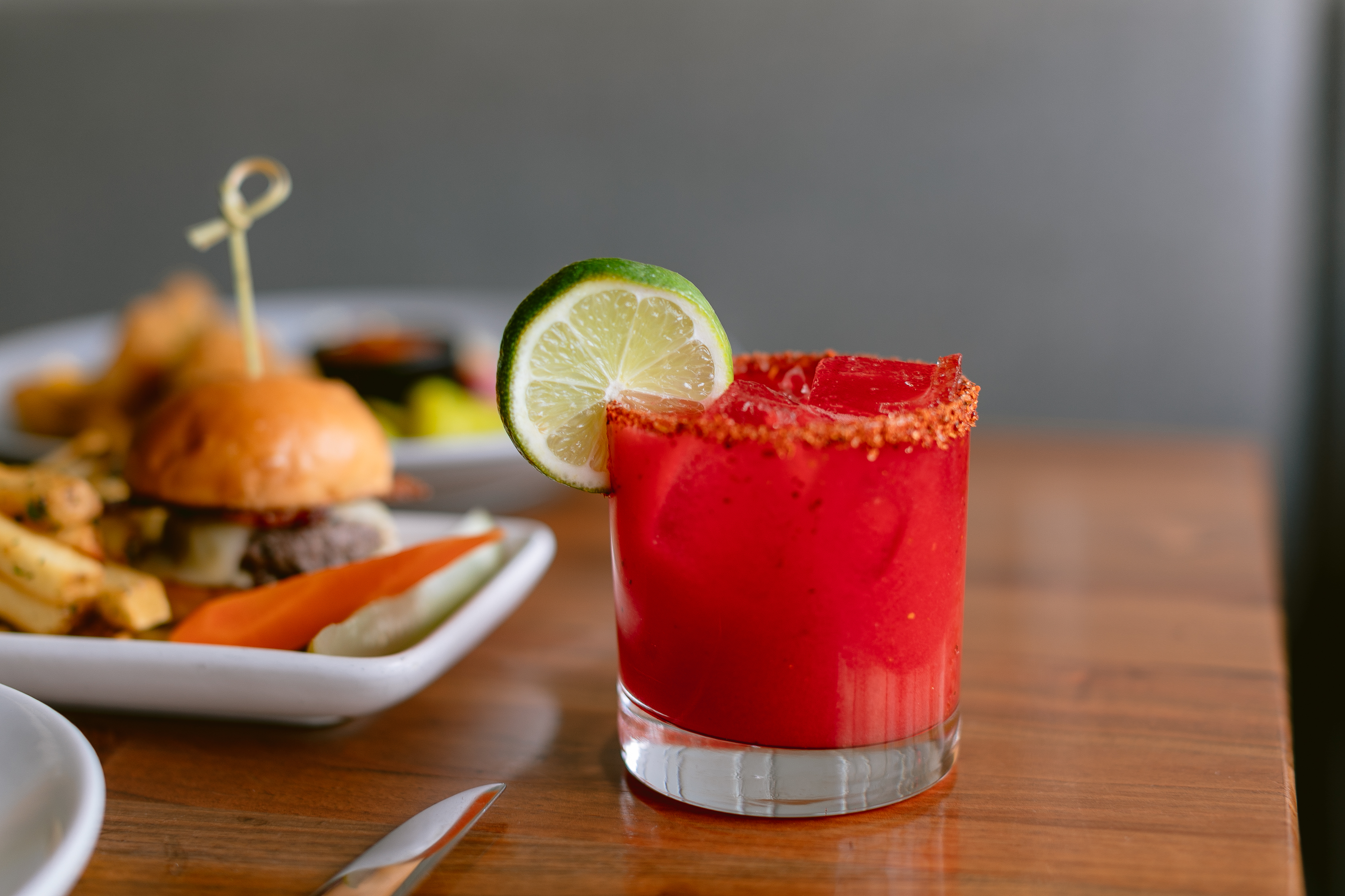 Photo of The Landsby. A vibrant red cocktail in a glass with a chili rim and a lime slice garnish sits on a wooden table, with a plate of a burger, fries, and pickles blurred in the background.