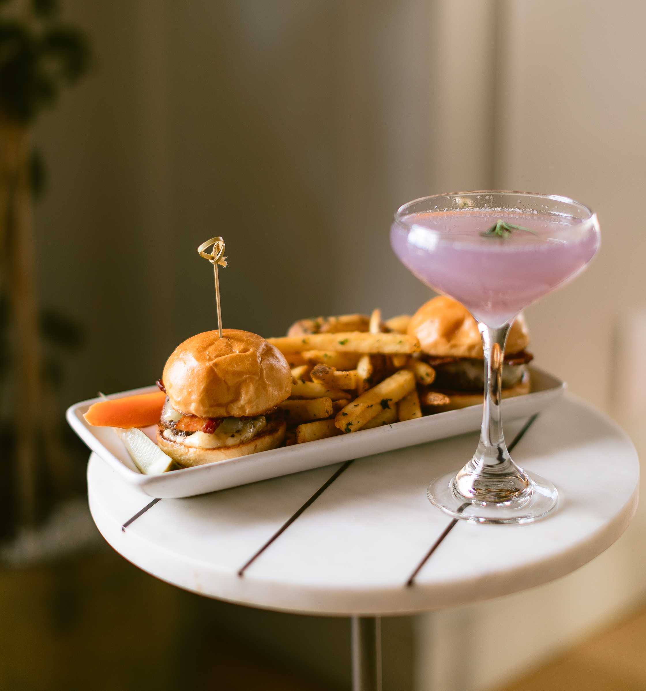 Photo of The Landsby. A rectangular plate with two sliders, a pile of seasoned fries, and pickle slices sits on a small round white table next to a purple cocktail in a coupe glass.