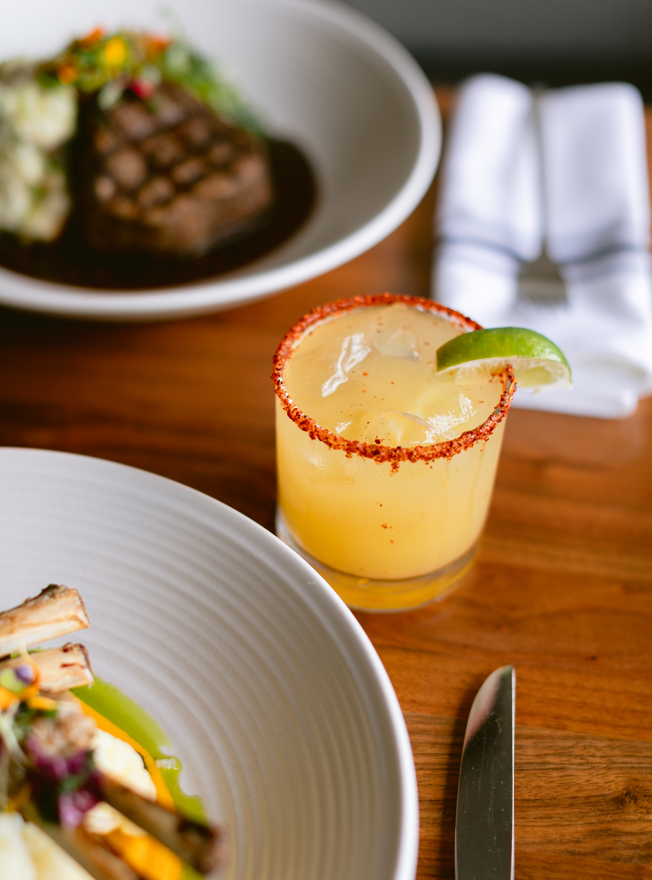 Photo of The Landsby. A cocktail with a chili-rimmed glass and lime wedge sits on a wooden table next to plated gourmet food, including ribs and a steak, with a folded napkin and knife nearby.