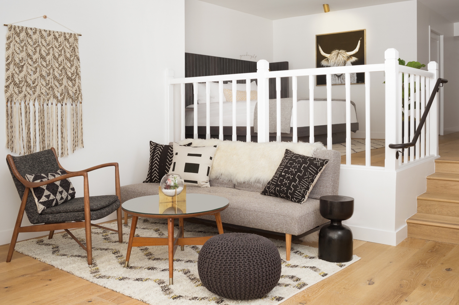 Photo of The Landsby. A modern living area with a gray sofa, patterned pillows, a wooden armchair, round glass coffee table, black side table, knitted pouf, and a white rug. A white railing separates the space from a bedroom above.