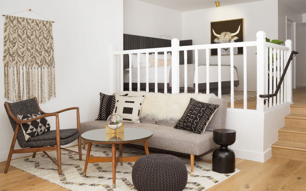 A modern Junior Suite living area features a gray sofa with black-and-white cushions, a glass coffee table, a wooden armchair, and a knitted pouf. Beyond the white railing, the bedroom offers a cozy bed. Decor includes wall hangings and artwork.