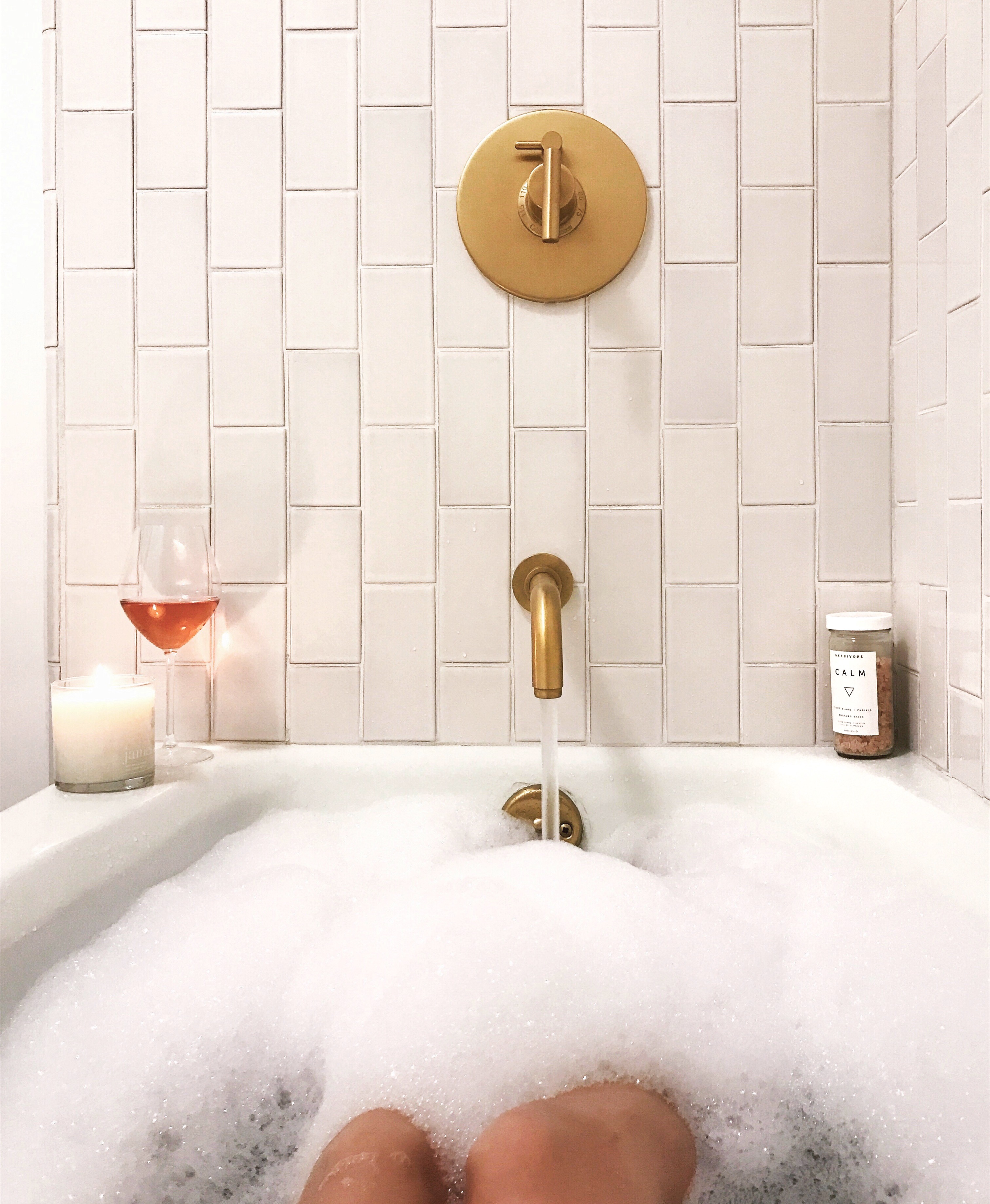 Photo of The Landsby. A person relaxes in a bubble-filled bathtub. A lit candle, a glass of rosé wine, and a container labeled calm are placed on the tub’s edge, with gold fixtures and white tile in the background.