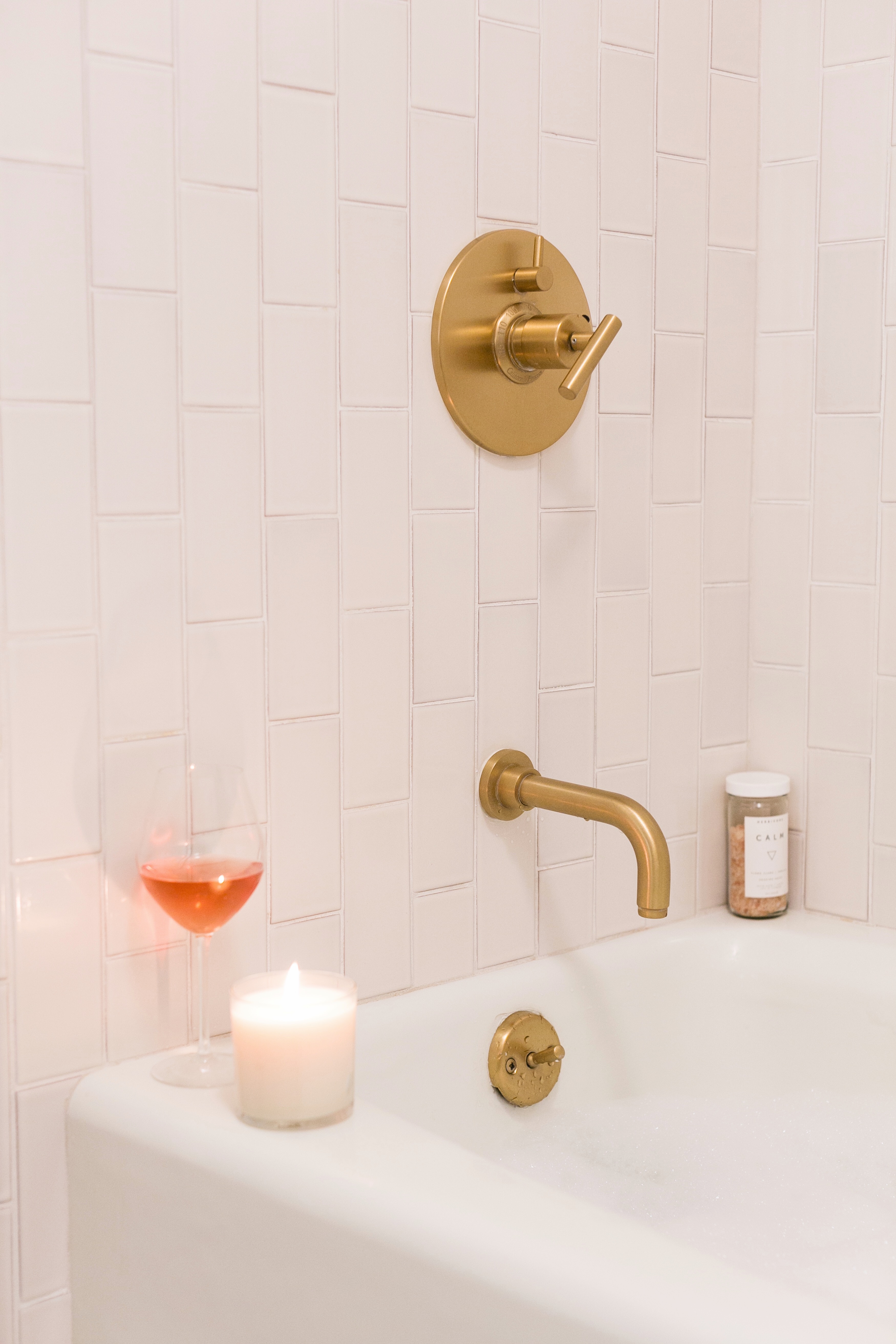 Photo of The Landsby. A bathtub with gold fixtures is filled with water. On the tub’s edge sit a lit white candle, a glass of rosé wine, and a small container. The white tile wall has a modern, vertical pattern.