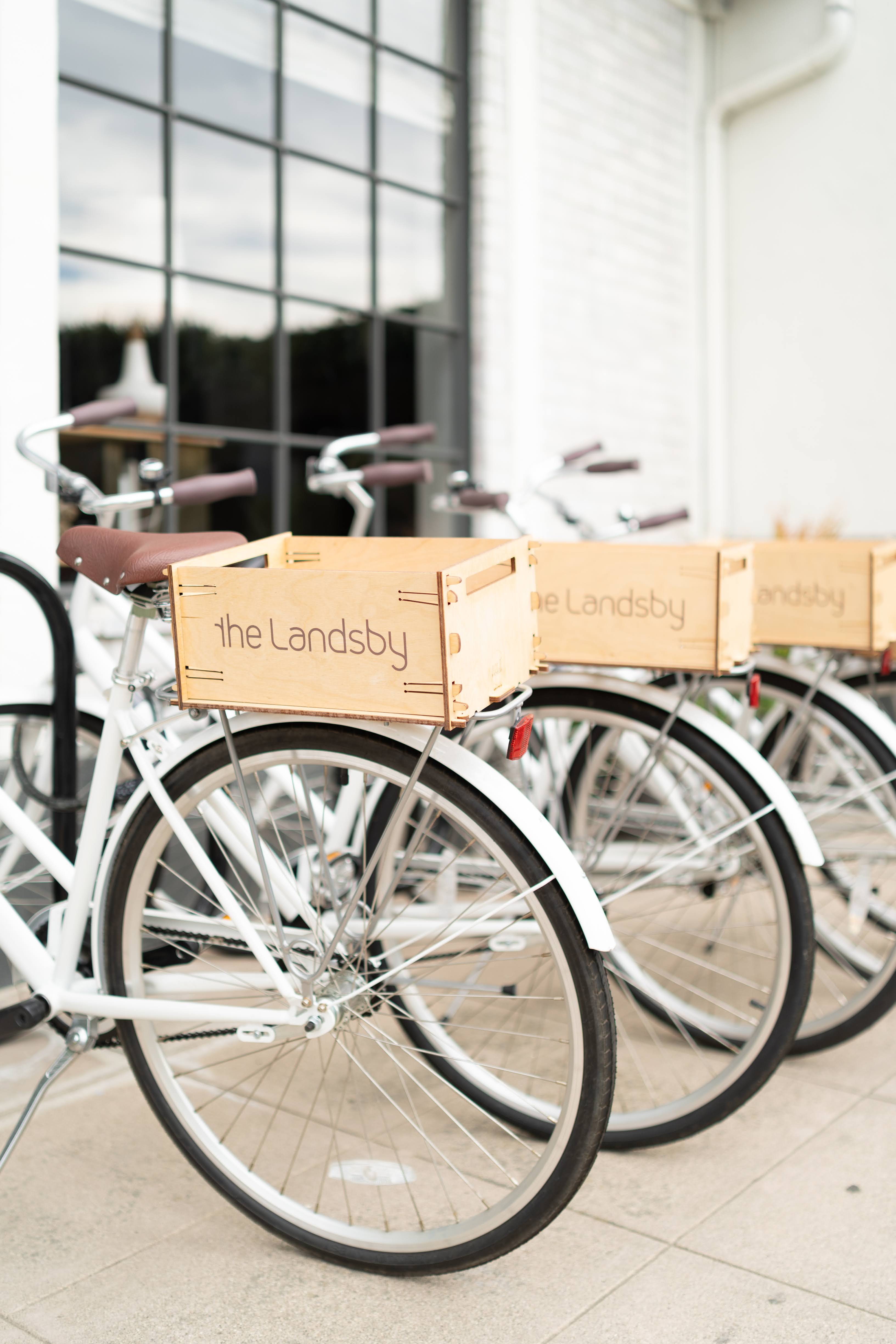 Photo of The Landsby. A row of white bicycles with wooden front baskets labeled the Landsby is parked on a light-colored sidewalk in front of a large window and white brick wall.