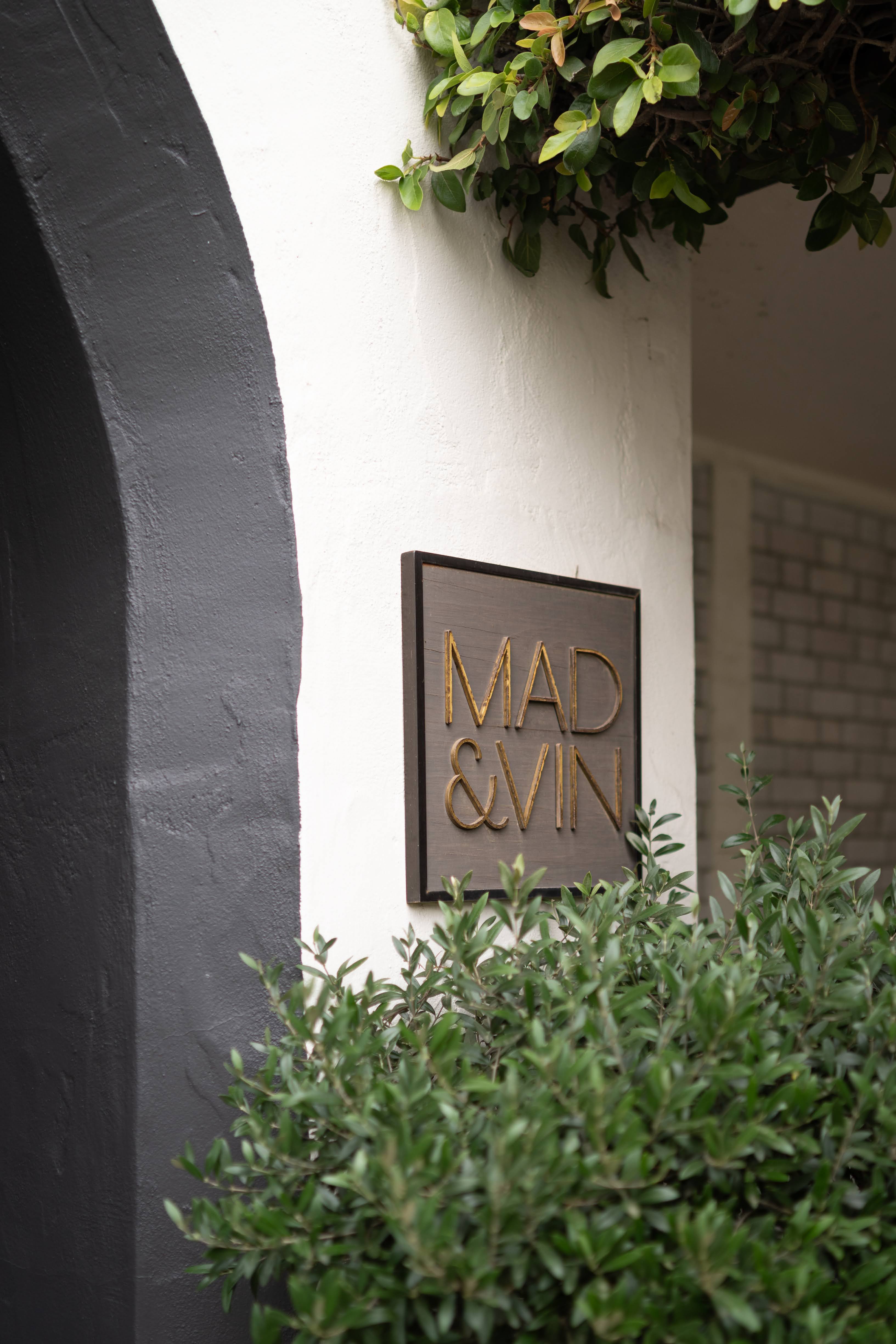 Photo of The Landsby. A metal sign reading MAD & VIN is mounted on a white wall next to a black archway, with green shrubs and overhanging leaves in the foreground.