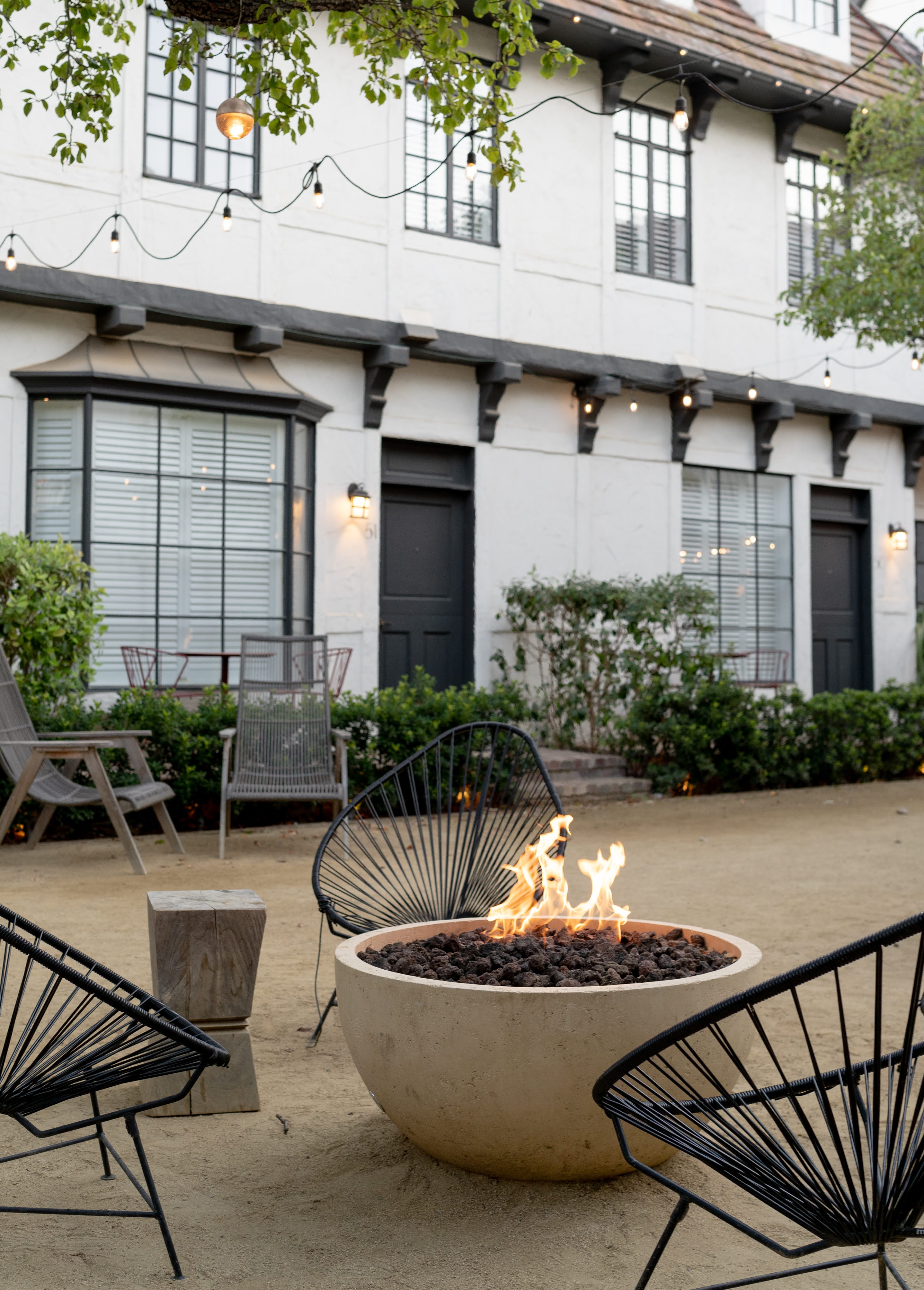 Photo of The Landsby. A round fire pit with flames is surrounded by modern black chairs in a sandy outdoor courtyard. String lights hang above, and a white building with black doors and windows is in the background.