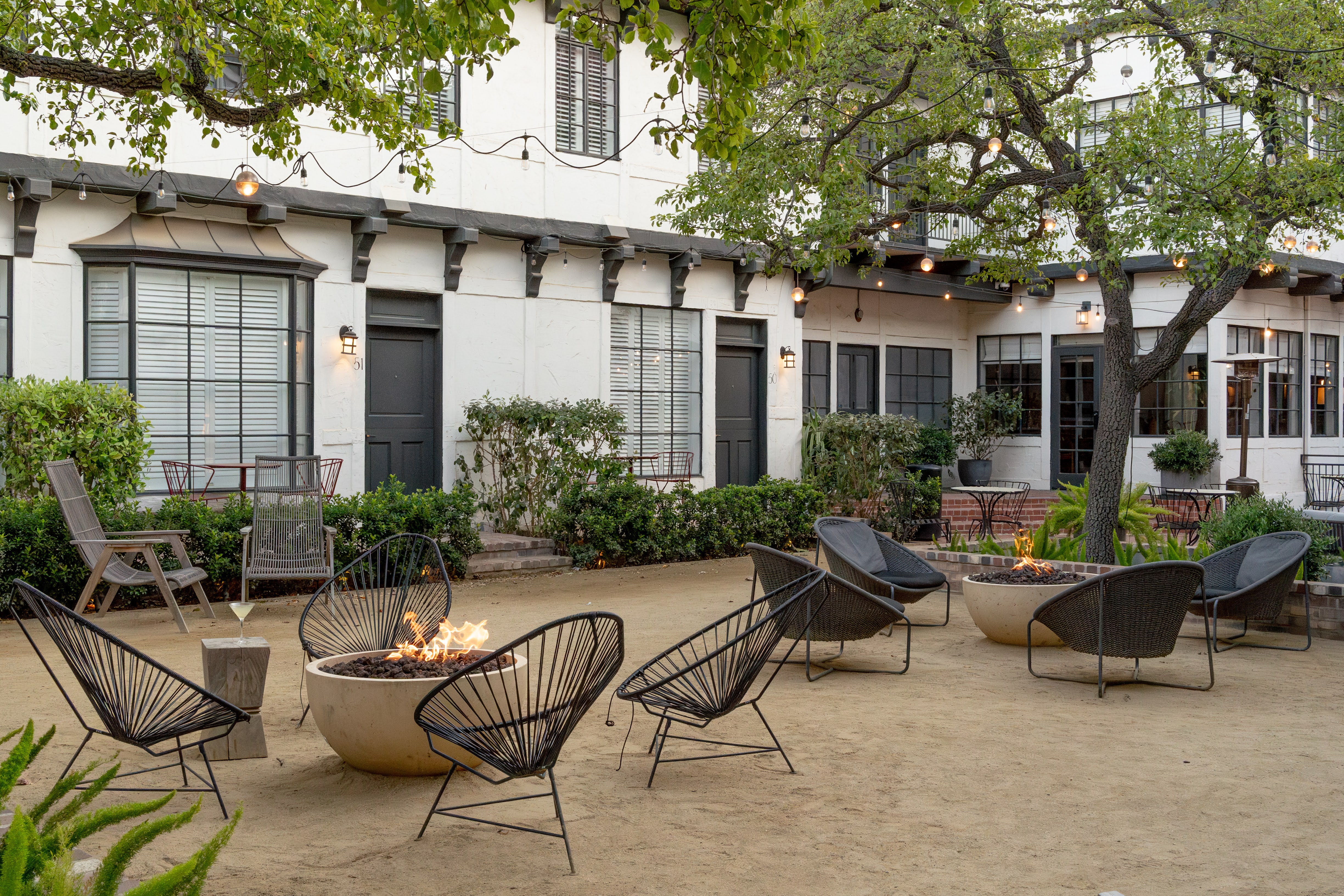 Photo of The Landsby. Outdoor patio area with modern black chairs and round fire pits, surrounded by greenery and trees, in front of a white building with multiple doors and windows. Warm, inviting ambiance with string lights overhead.
