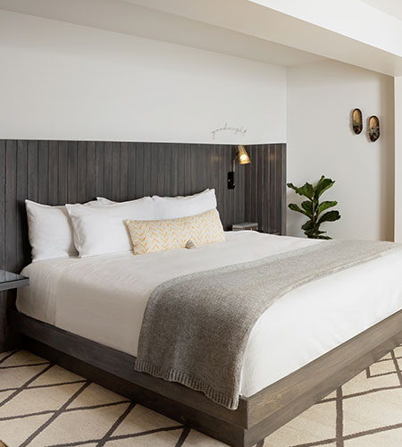 A modern, minimal bedroom with a large bed featuring white linens, a gray throw blanket, and a chevron accent pillow. The room has a dark wood headboard, plant, wall art, and a patterned rug.