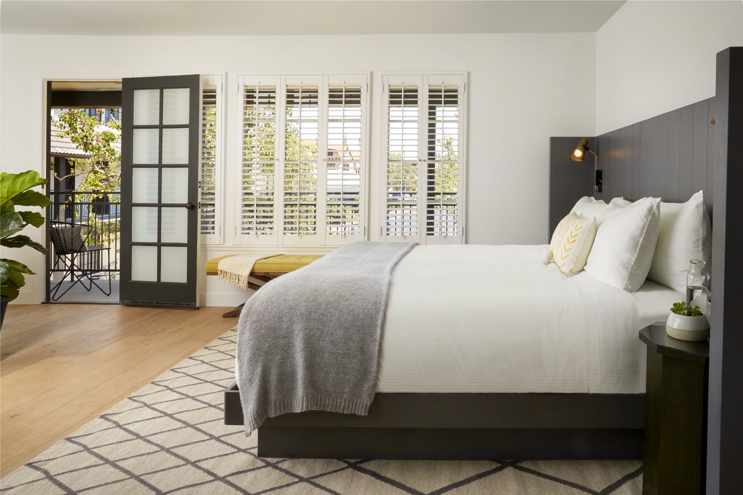 Photo of The Landsby. A bright, modern bedroom with a large bed, white bedding, gray throw, and patterned rug. There are glass doors open to a balcony with outdoor seating, and plantation shutters let in natural light.