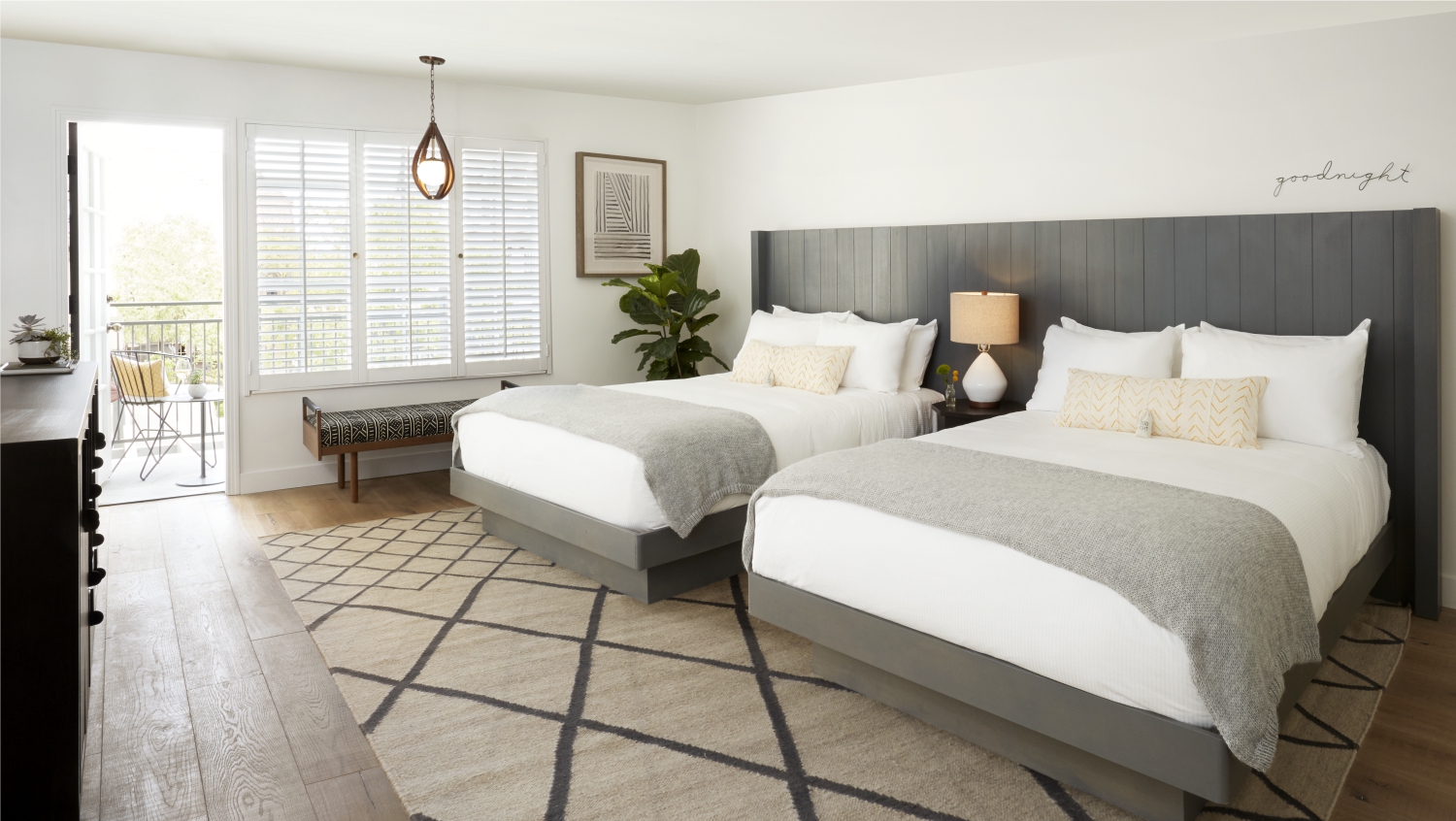 Photo of The Landsby. A bright, modern hotel room with two large beds, gray headboards, white linens, a geometric rug, a lamp, a plant, a bench by the window, and a balcony visible through open glass doors.
