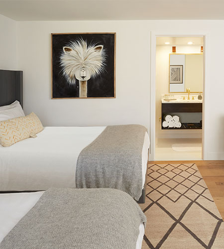 A modern hotel room with two beds covered in gray blankets, a geometric rug, and a large framed llama portrait above the beds. An open doorway reveals a well-lit bathroom with towels and a sink.