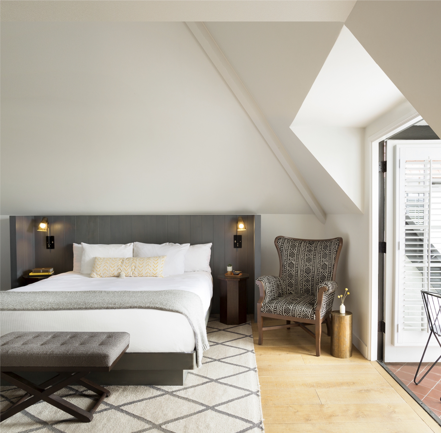 Photo of The Landsby. A cozy bedroom with a large bed, patterned armchair, wooden bench, bedside tables, and wall lamps. Natural light enters through a window with white shutters, and a geometric rug covers the wooden floor.