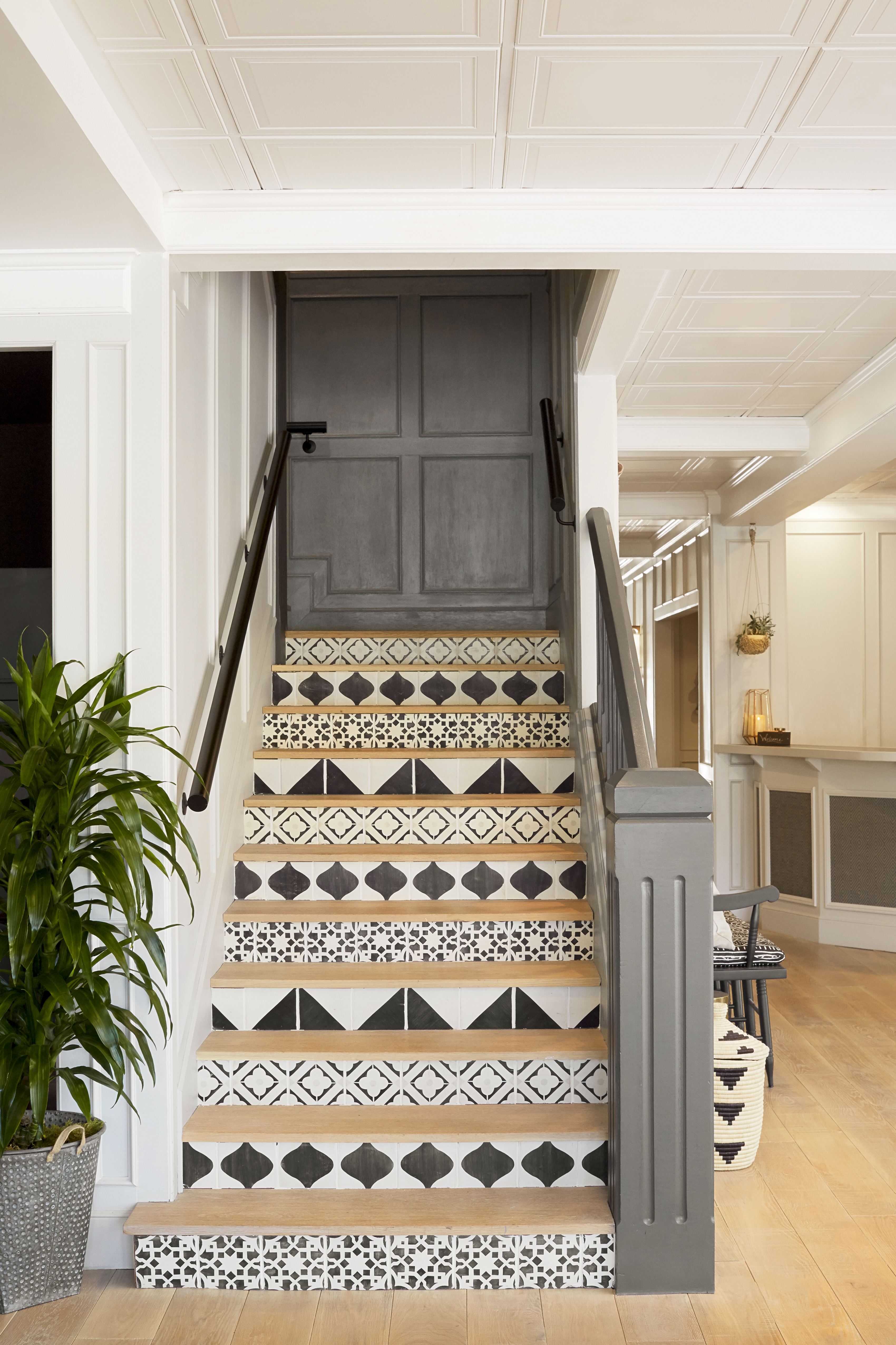 Photo of The Landsby. A staircase with patterned black and white tiles on the risers, a dark handrail, and a gray door at the top. A potted plant is on the left, and the space has light walls and a wooden floor.