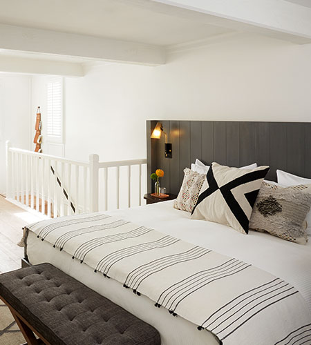 A bright, modern bedroom with a white bed, striped throw blanket, patterned pillows, dark headboard, wall-mounted reading light, tufted bench, and white railing along the staircase.
