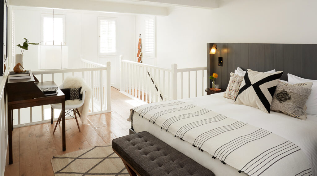 A bright, modern bedroom with a neatly made bed, geometric pillows, a striped blanket, a wooden desk with a chair, and natural light streaming in through shuttered windows and white railing.