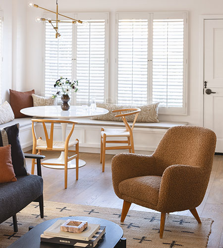 A cozy, modern living and dining area with a tan armchair, round white dining table, wooden chairs, built-in bench seating, and large windows with white shutters. Neutral tones and minimalist decor create a warm atmosphere.
