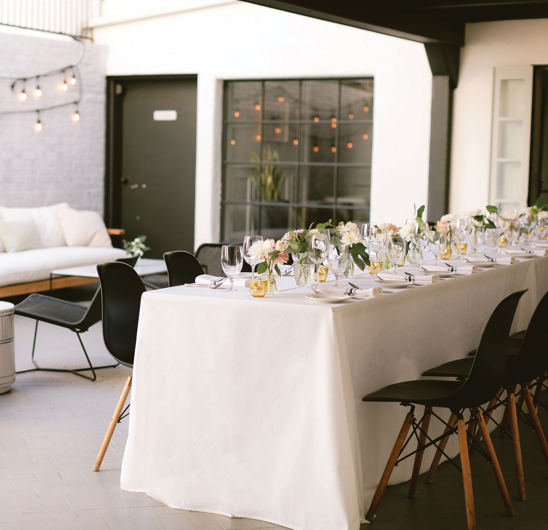 A long table with a white tablecloth is set for a formal meal with glassware, plates, and floral centerpieces. Black chairs line both sides, and string lights create a cozy atmosphere in a modern indoor-outdoor space.