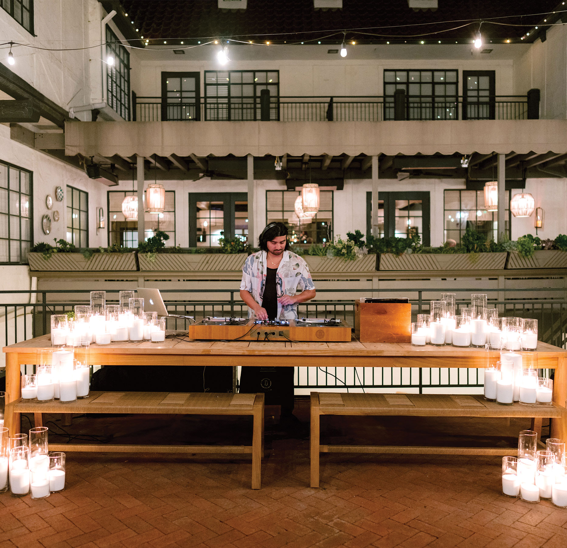 A DJ stands behind a wooden table with DJ equipment and many lit candles in glass holders, set in a courtyard with string lights and a building with balconies and large windows in the background.
