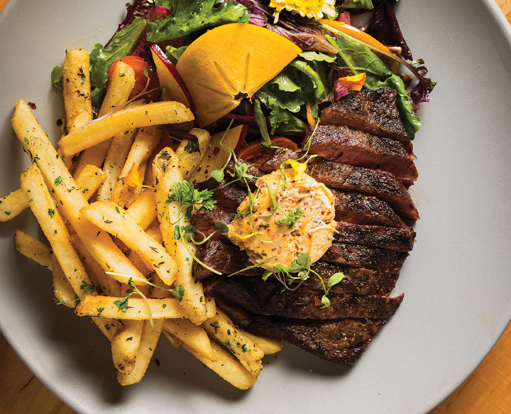 where to eat in Solvang A plate with grilled steak topped with herb butter, golden French fries sprinkled with herbs, and a mixed green salad featuring a tomato wedge and yellow edible flower—perfect for those seeking Top Solvang Dining.