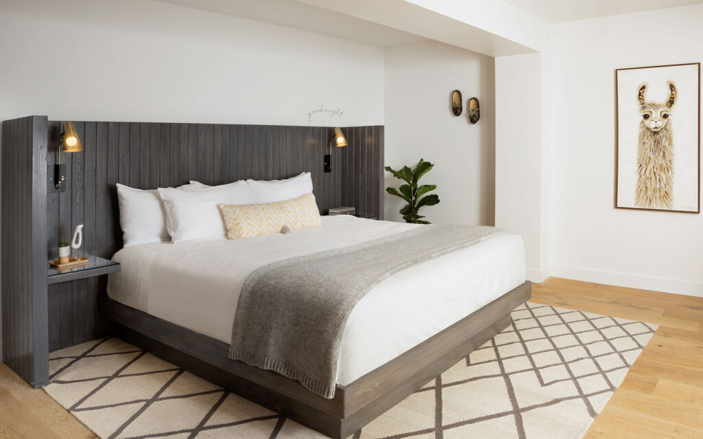 A modern Village Suite bedroom with a large bed, wooden headboard, white bedding, and a gray throw. Wall lamps, a geometric-patterned rug, a potted plant, and framed llama artwork enhance the cozy white-walled space.