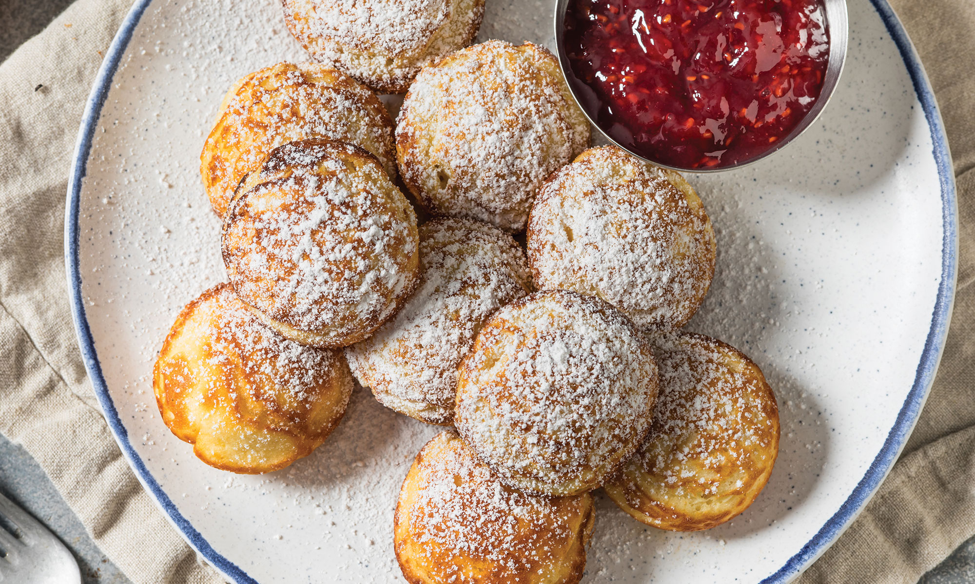 A plate of round, golden-brown pastries dusted with powdered sugar, served with a small bowl of red fruit jam on the side—a delicious treat to enjoy while exploring Things To Do In Solvang.