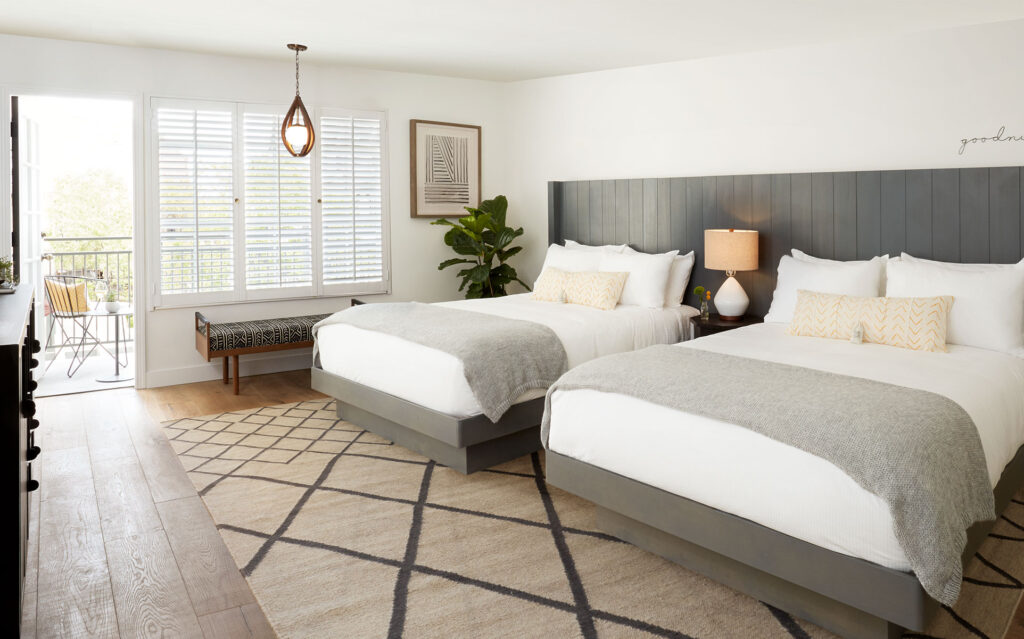 Bright hotel room with two queen beds, chic gray headboard, white bedding, cozy throws, and a large window with shutters—one of the inviting places to stay in Solvang featuring a small balcony overlooking the charming surroundings.