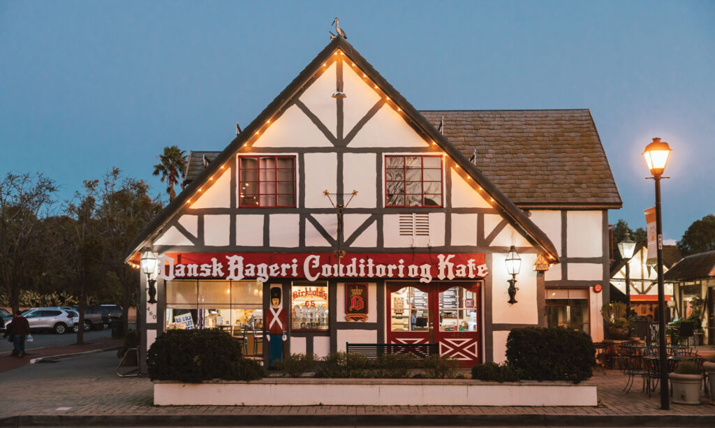 A half-timbered bakery with a steep roof and lit signs reading Dansk Bageri Conditoring Kafé stands on a street at dusk—one of the charming things to do in Solvang, its warm lights inviting visitors as a few parked cars rest nearby.