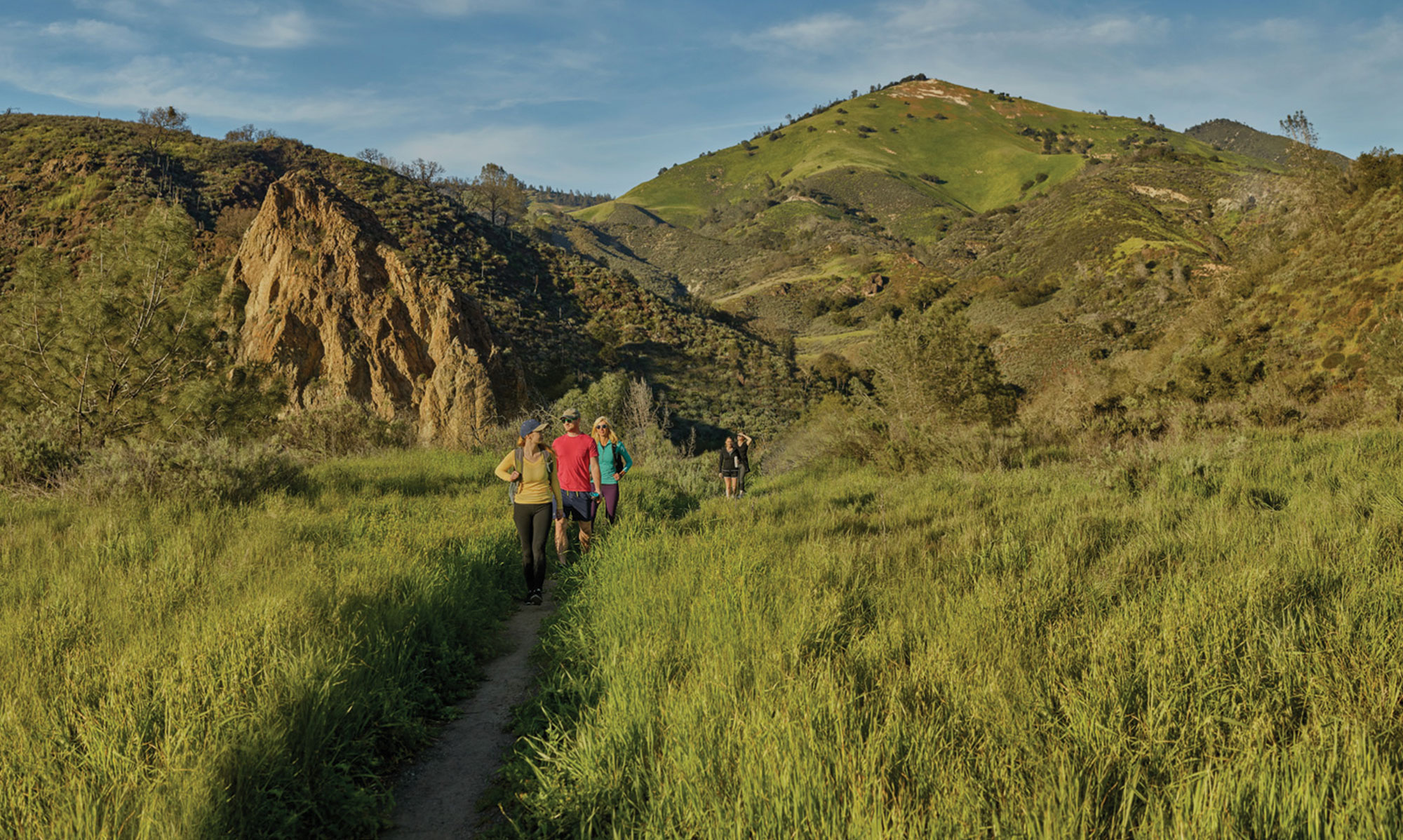 A group of hikers walk along a narrow trail through green grass and rolling hills under a partly cloudy sky, with mountains in the background—one of the many scenic things to do in Solvang.