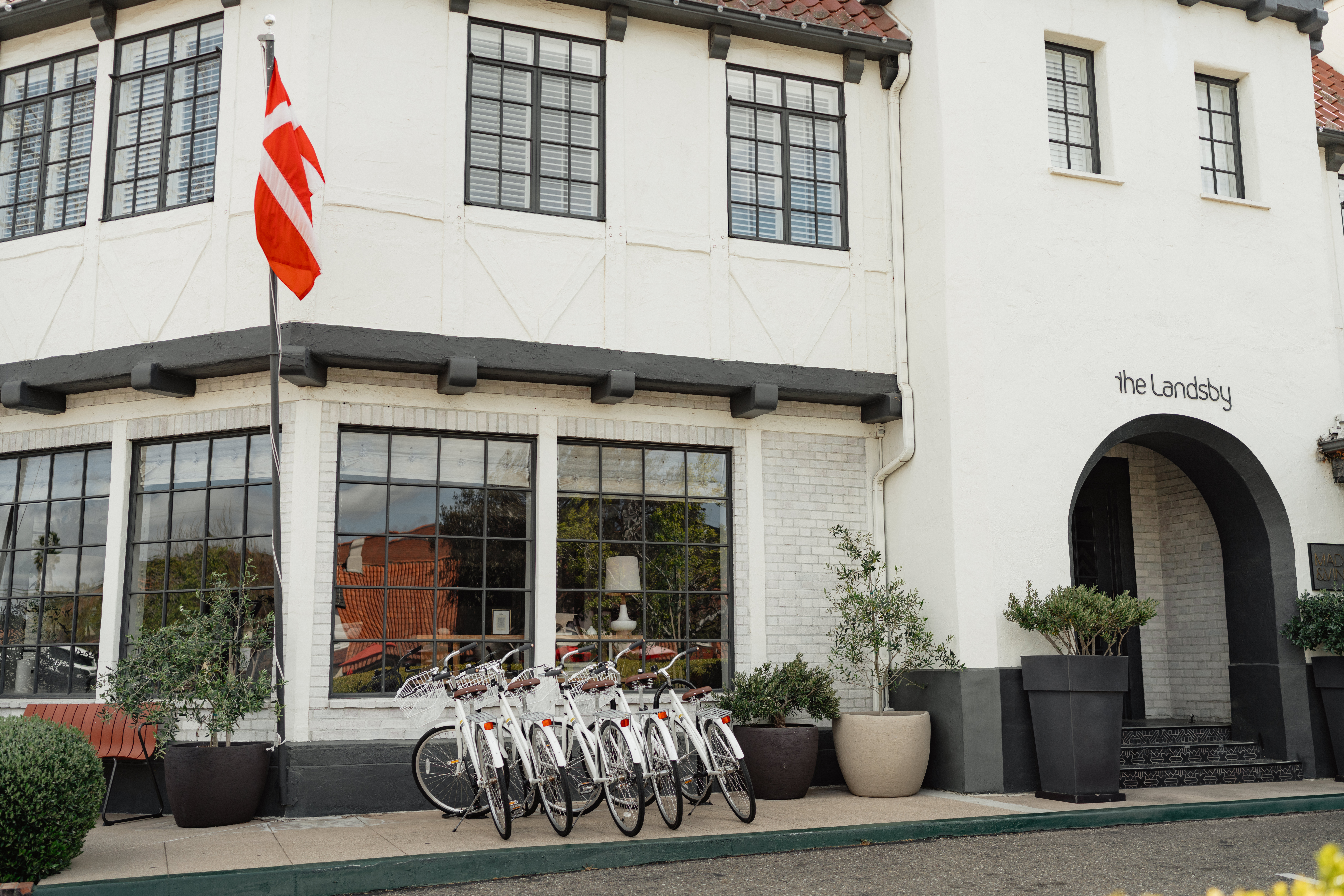 Photo of The Landsby. A row of white bicycles is parked outside a white building with large windows, potted plants, a Danish flag, and a sign that reads “the Landsby.”.