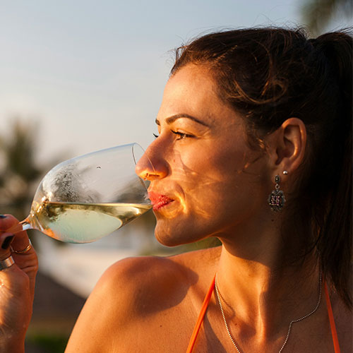 A woman with dark hair and earrings is sipping white wine from a glass outdoors, bathed in warm sunlight, with a blurred background of trees and sky.