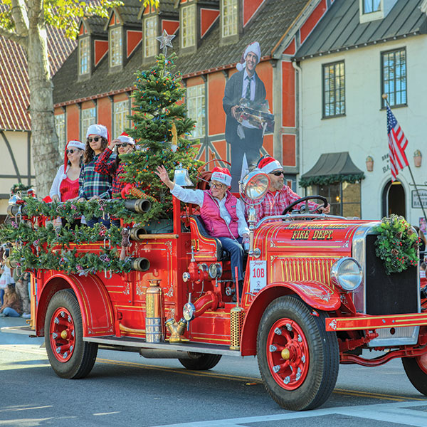 Best Christmas Town in California A vintage red fire truck, decorated with Christmas garlands and a tree, carries people in festive attire and Santa hats during a parade. In the background, a large cardboard cutout of a man with a tray promotes Press & Media coverage.